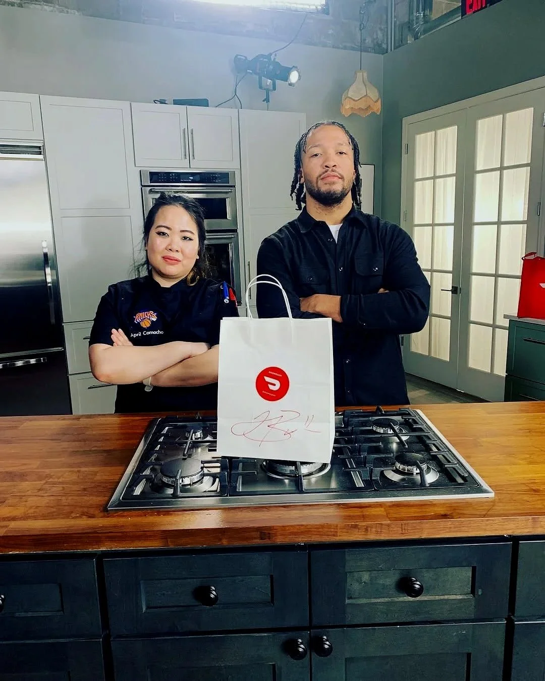 Two individuals standing behind a kitchen island with a gas stove, one woman and one man, with the woman crossing her arms and the man with his arms crossed. A paper bag with a red logo and a signature is on the counter. The woman is wearing a dark chef's jacket with a patch that says 'Knicks' and the name 'April Comacho.' The man is wearing a black shirt. The background has kitchen cabinets, a double oven, a refrigerator, and a glass door, with lighting equipment visible.