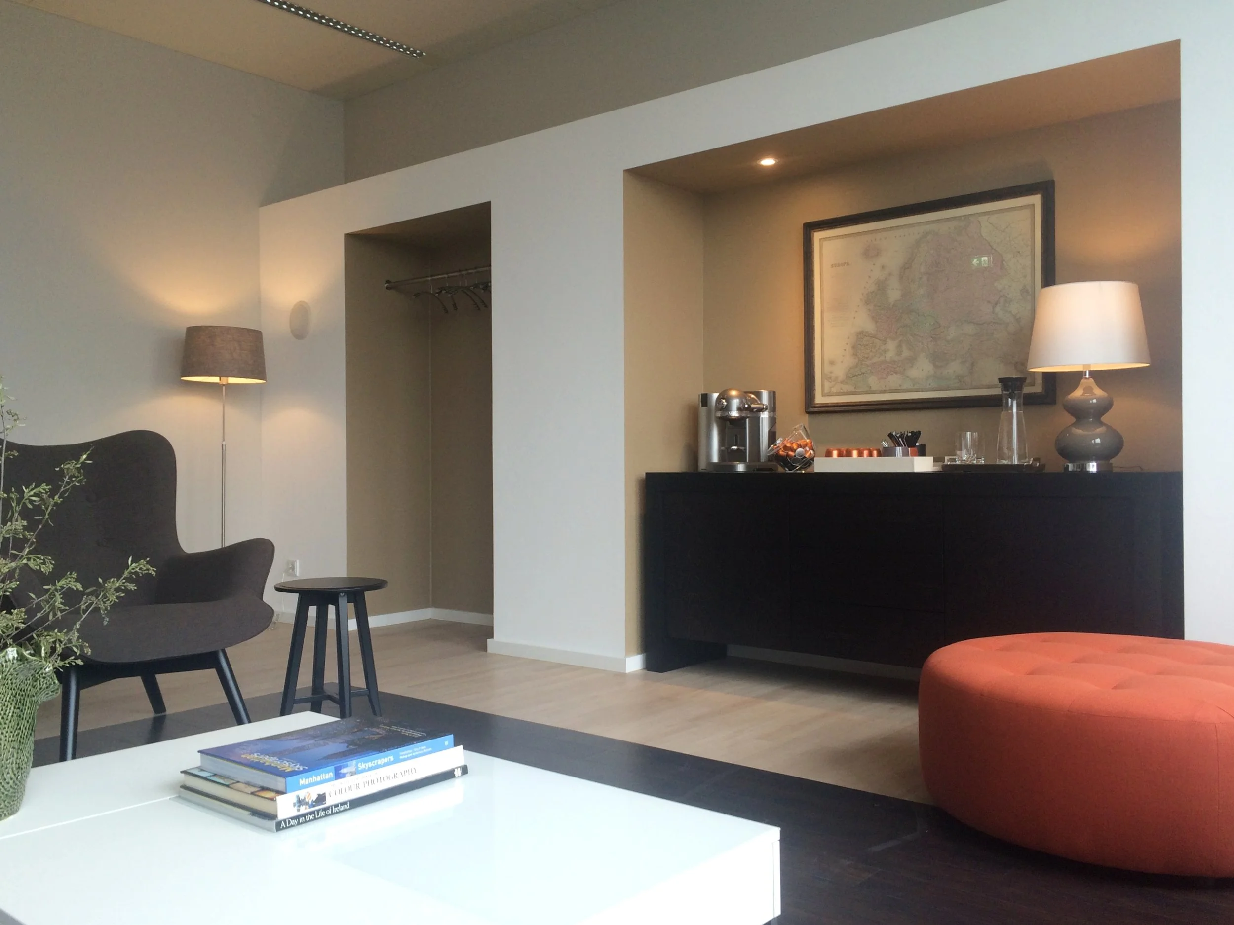 Innenaufnahme eines modernen Wohnzimmers mit dunklem Sofa, kleinen Beistelltischen, Stehlampen, einem Schwarzsideboard mit einer Kaffeemaschine und anderen Getränkebehältern, sowie einer Karte an der Wand, einem großen roten Sitzkissen und Büchern au