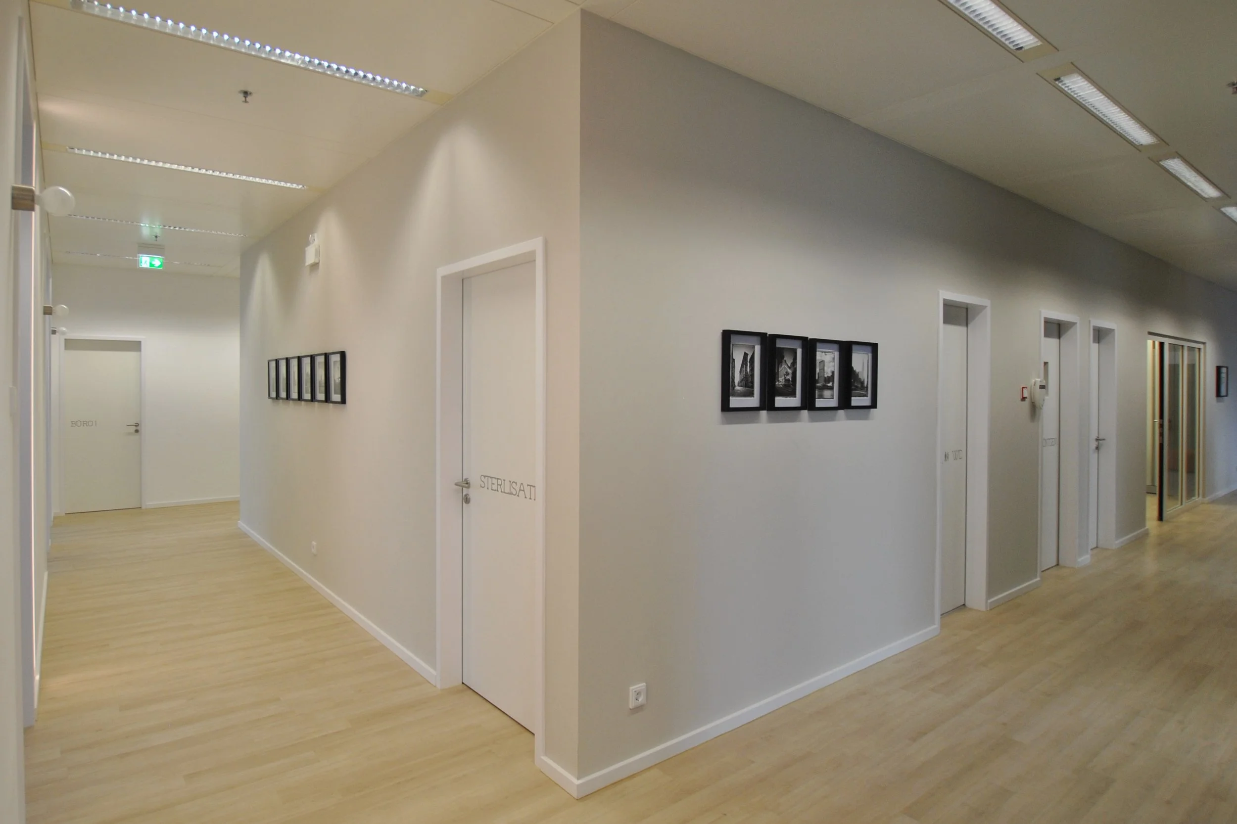 Innenansicht eines Flures mit weißen Wänden, mehreren Türen, Wanddekorationen, und Holzfußboden in einem modernen Gebäude