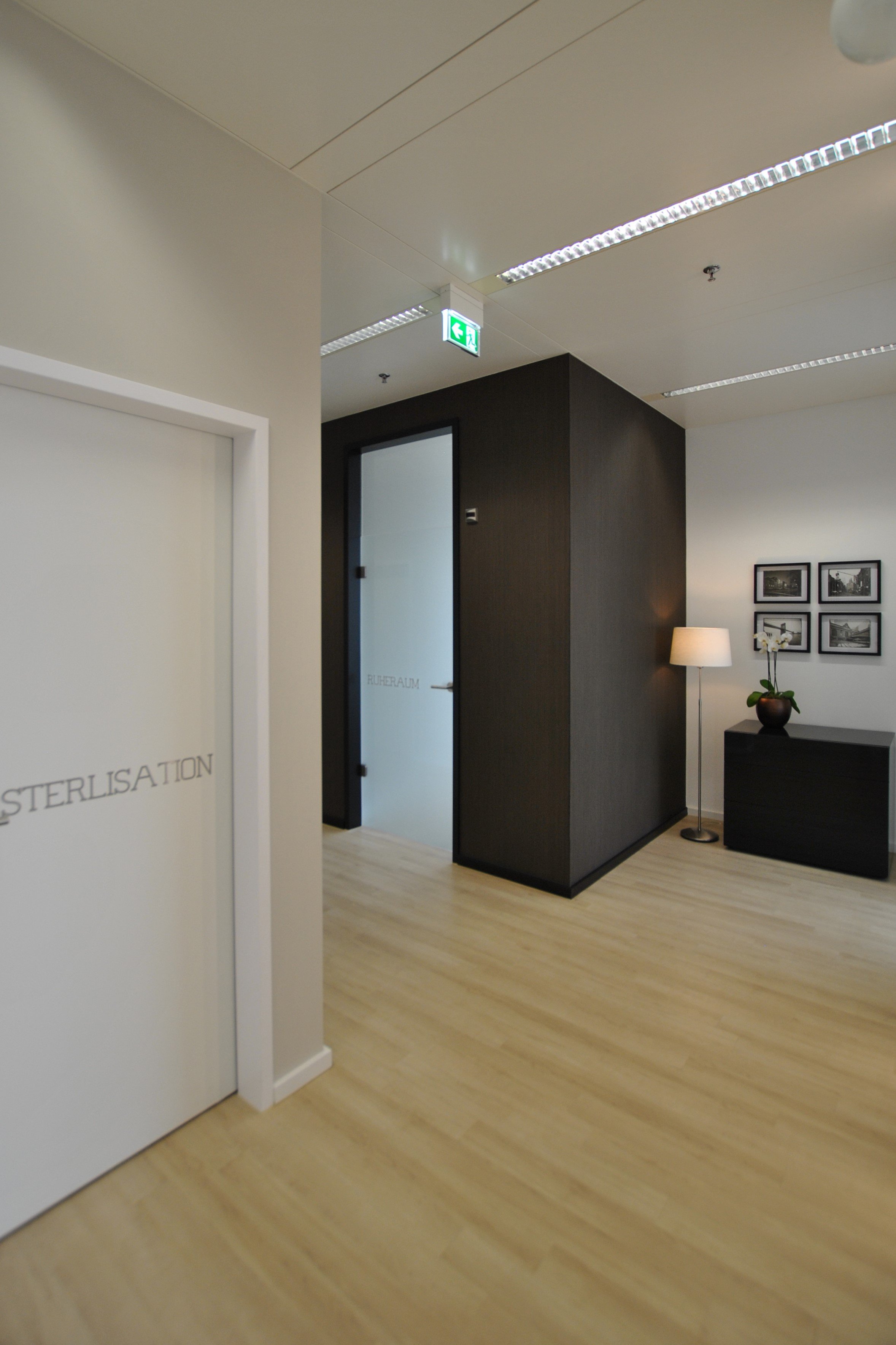 Ein Flur in einem modernen Gebäude mit Holzfußboden, weißen und schwarzen Wänden, einem kleinen Beistelltisch mit einer Lampe, einer Vase und einer Blume, sowie vier Schwarz-Weiß-Fotos an der Wand.