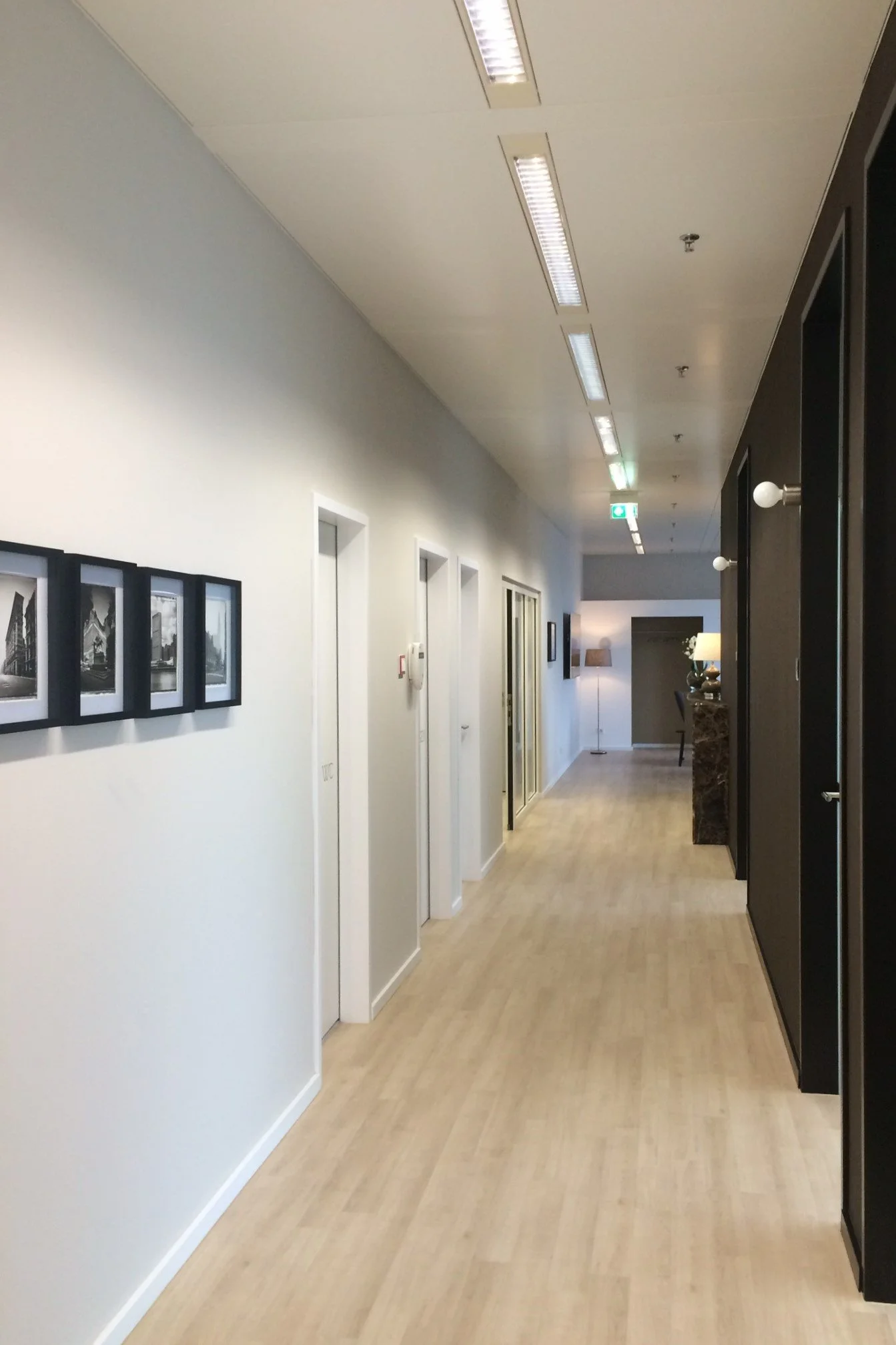 Ein heller Flur in der Praxis Dr. Hakimi + Kollegen in einem modernen Gebäude mit weißen Wänden, Holzfußboden und Deckenleuchten. Es gibt Bilder an der Wand und Türen auf beiden Seiten, einige mit Glastüren im Hintergrund.