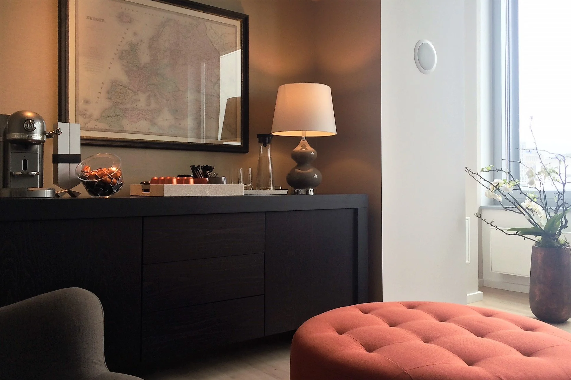 Innenraum mit Schwarzer Kommode, Tischlampe, Landkarte an der Wand, Kaffeebereiter, Glas mit Kaffee-Kapseln, Blumen in Vase, Sessel, roter Sitzhocker, Fenster mit Blick nach draußen, entspannte Wohnzimmerdekoration.