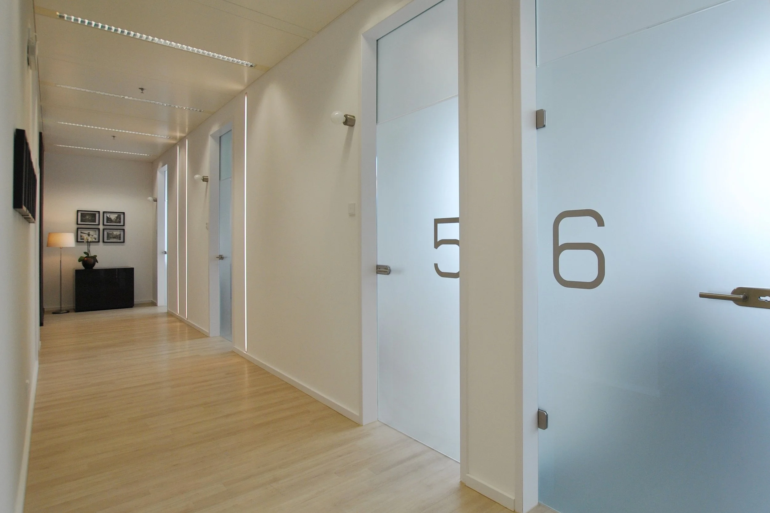 Heller Flur im modernen Gebäude mit weißen Wänden, Holzboden, Türen mit großen Nummern 5 und 6, und einer kleinen Ecke mit Lampen und Bildern.