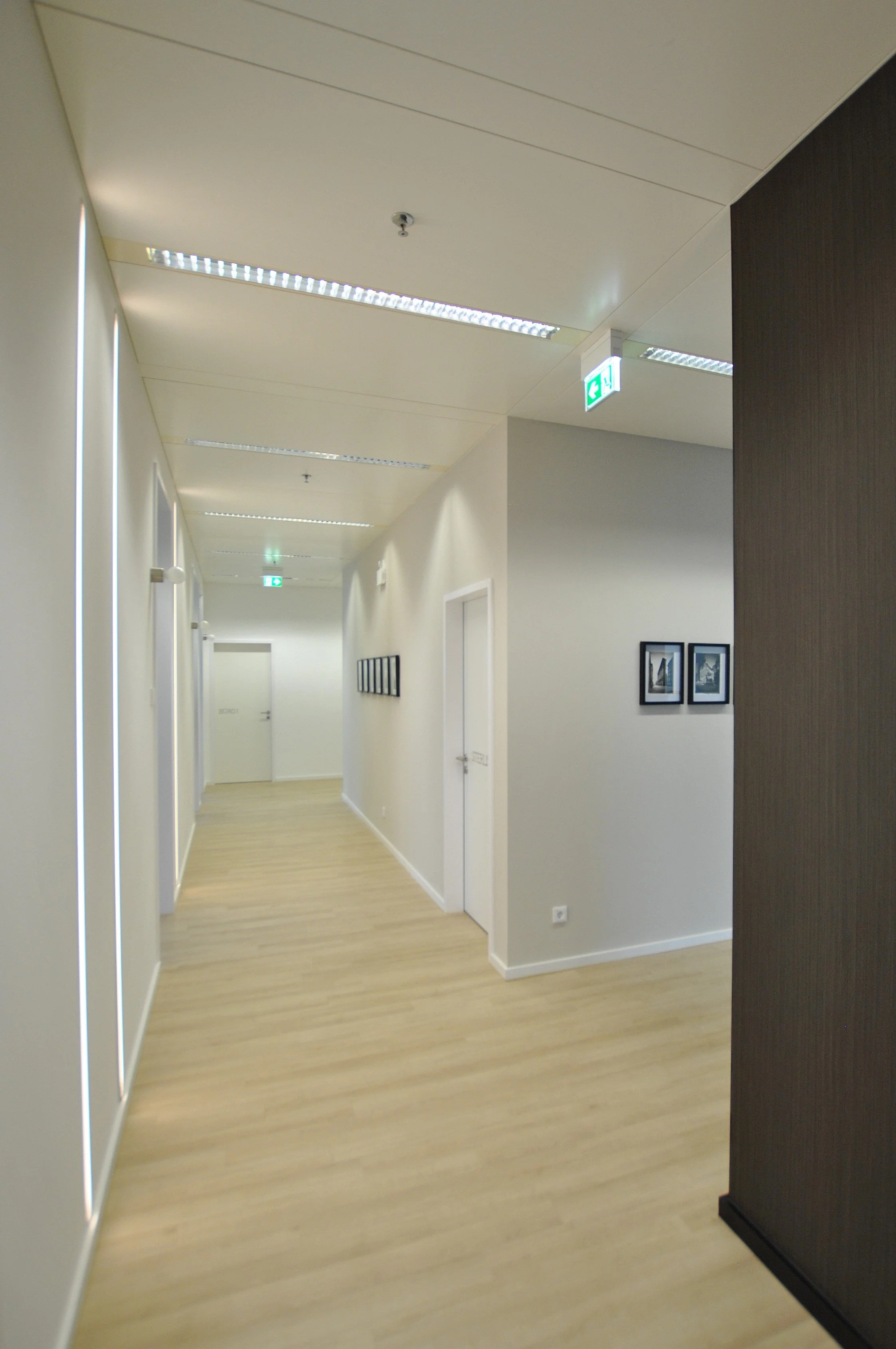 Ein heller Flur in einem modernen Gebäude mit weißen Wänden, Holzboden, mehreren ausgestellten Bildern und beleuchteten Deckenleuchten, sowie einem grünen Notausgangsschild.