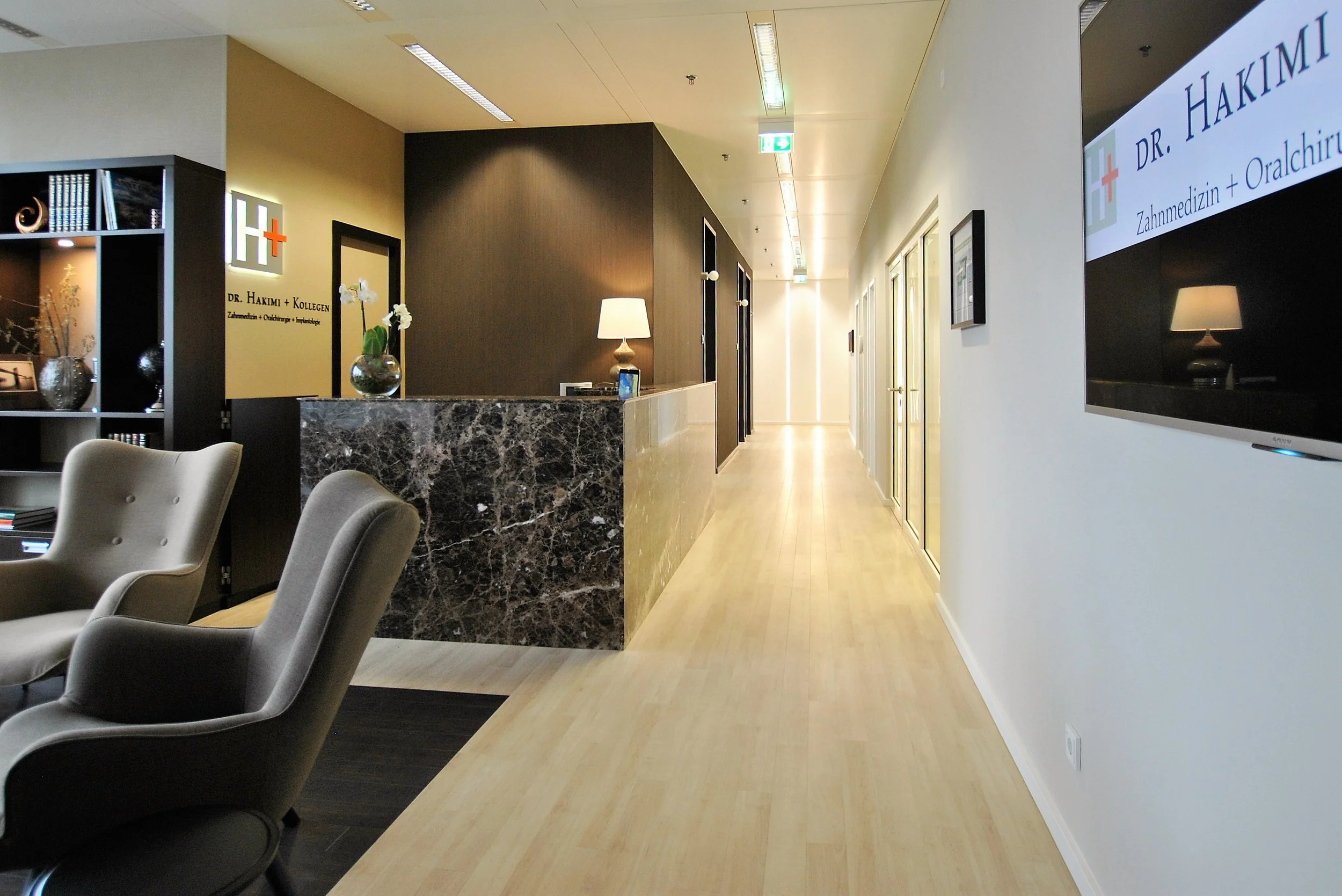 Empfangsbereich einer Zahnarztpraxis mit Stehlampen, Bücherregal, Sessel, Empfangstheke aus Marmor und einem Wandbild mit der Aufschrift 'Dr. Hakimi + Kollegen'.