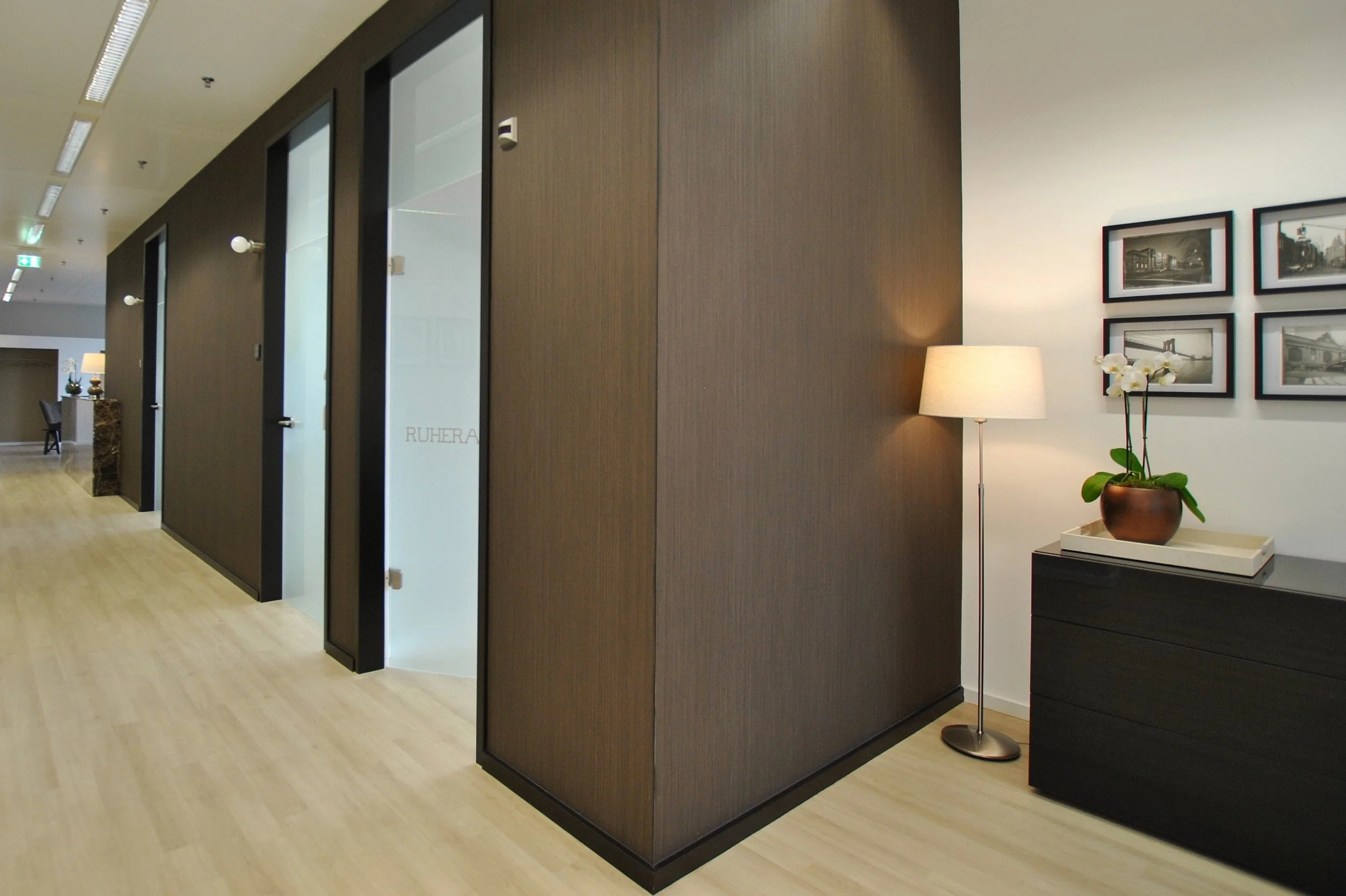 Moderne Hotellobby mit Holzfußboden, dunklen Wandpaneelen, einem Sideboard mit einer Orchidee und einem Stehlampe, sowie Fotos an der Wand.