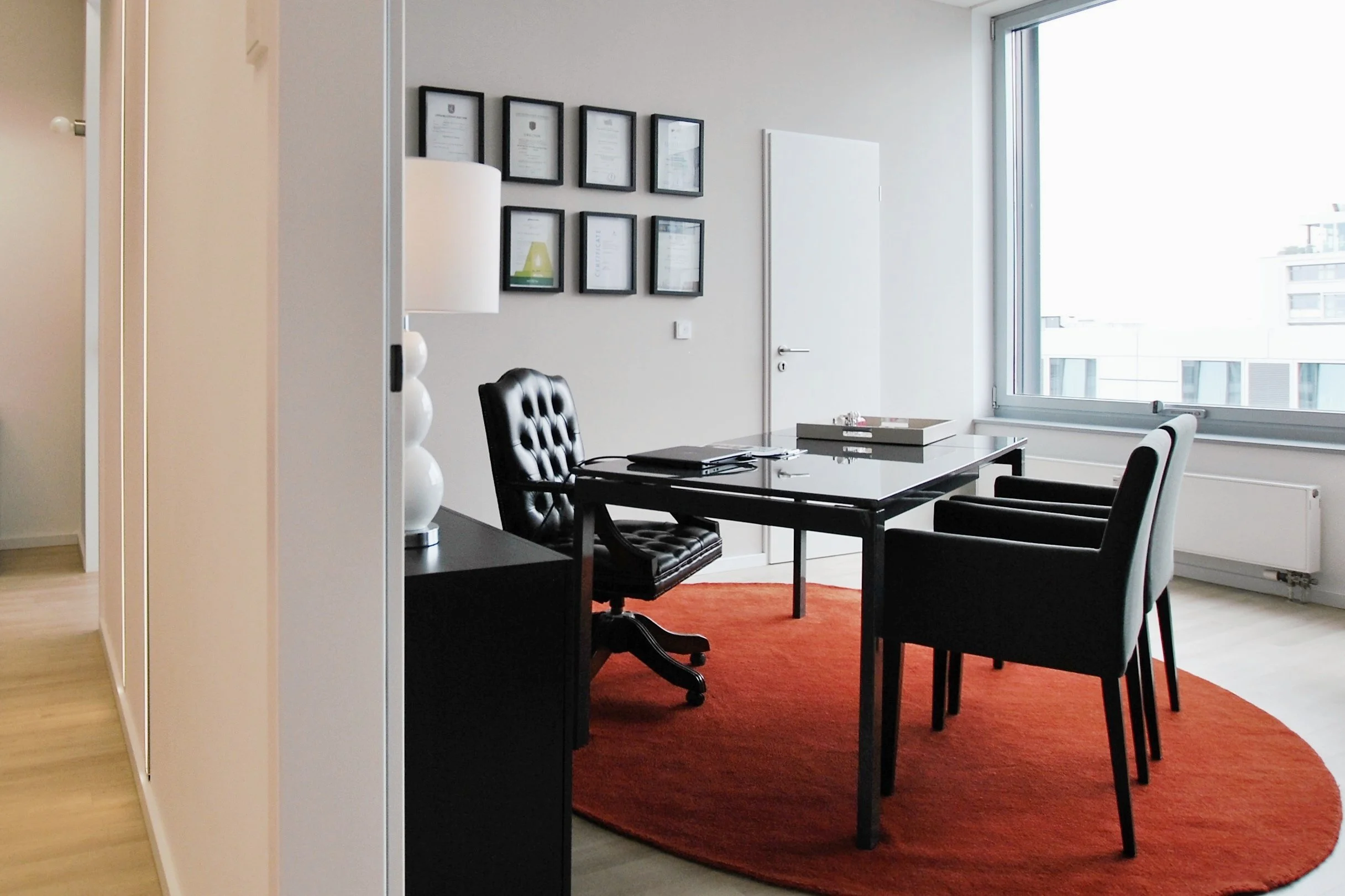 Ein modernes Büro mit schwarzem Tisch, drei schwarzen Stühlen, einem schwarzen Bürostuhl, einem weißen Lampenschirm auf einem schwarzen Sideboard, gerahmten Zertifikaten an der Wand, großem Fenster mit Blick auf andere Gebäude, und einem roten runden