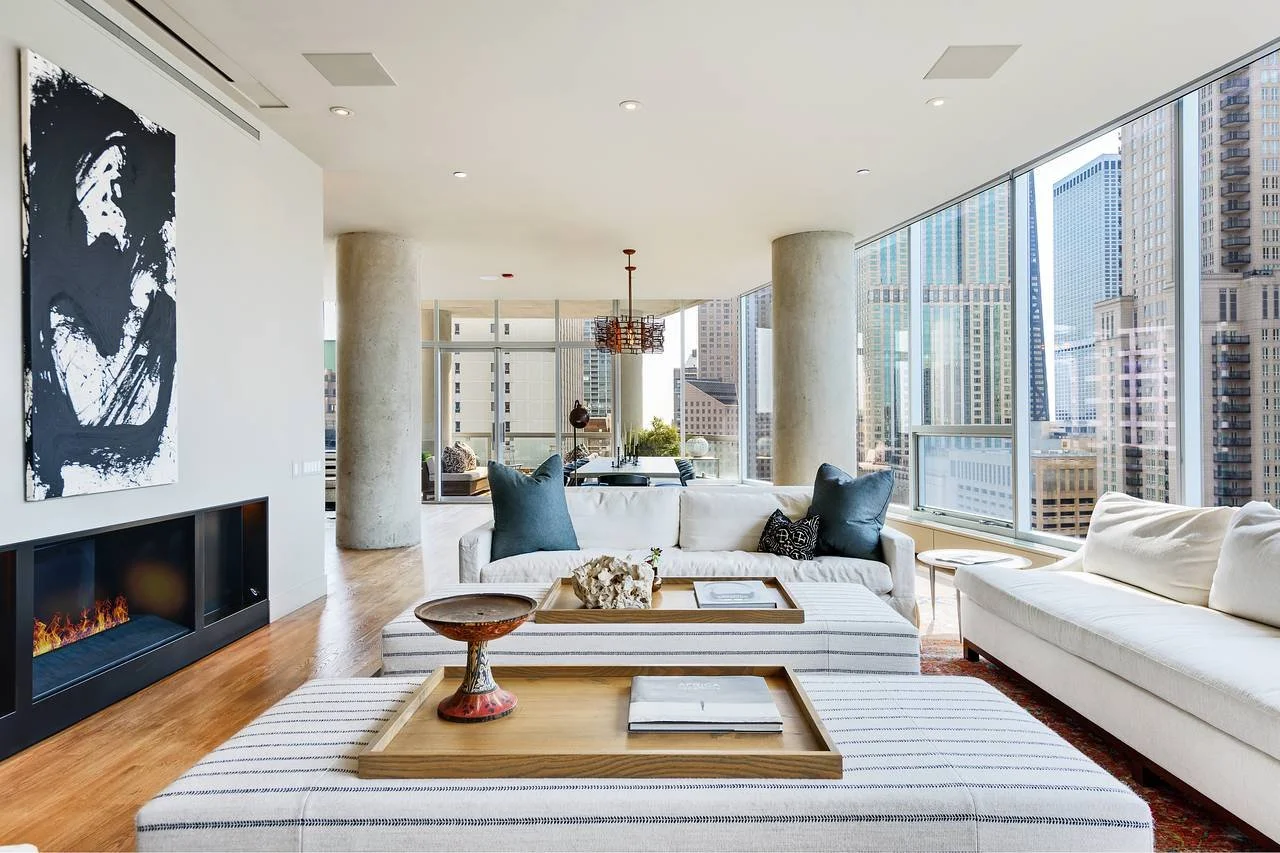 Modern living room with large windows overlooking city skyline, white sofas with dark pillows, wooden coffee tables, a painting on the wall, and a fireplace.