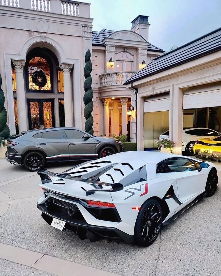 A white Lamborghini sports car with a black spoiler parked in front of a luxury house, with a sleek black SUV and other cars nearby.