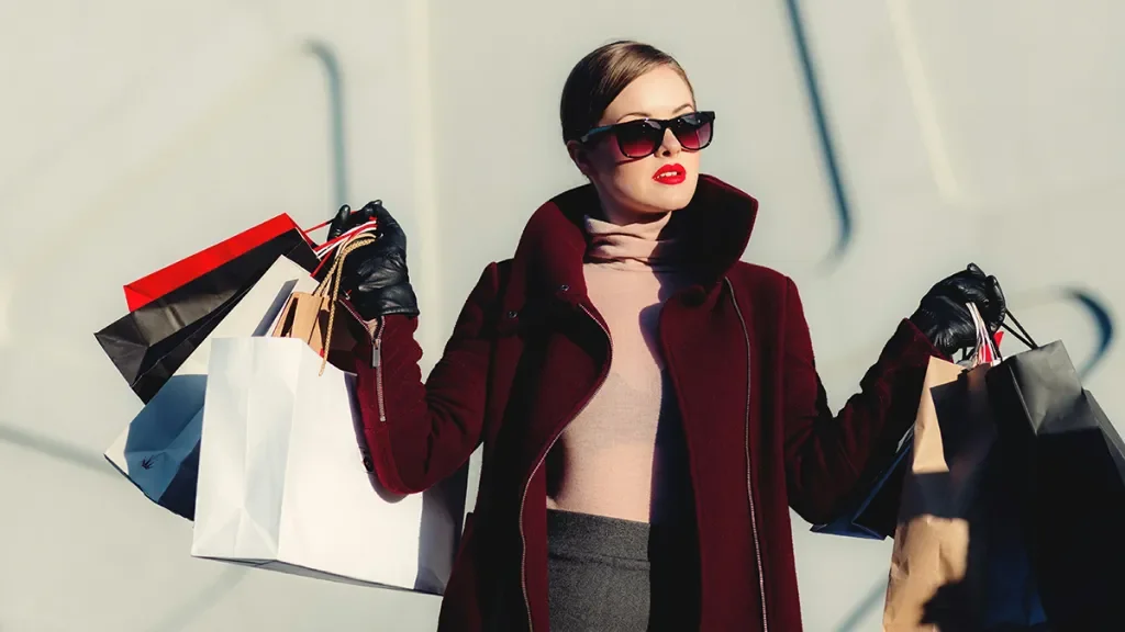 A woman wearing sunglasses, a maroon coat, and leather gloves holding shopping bags.