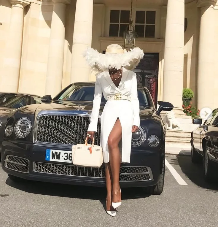 A woman dressed in a stylish white outfit with a front slit, wearing a large wide-brimmed hat with fluffy details, stands in front of a luxury black car, likely a Bentley, in front of an elegant building.