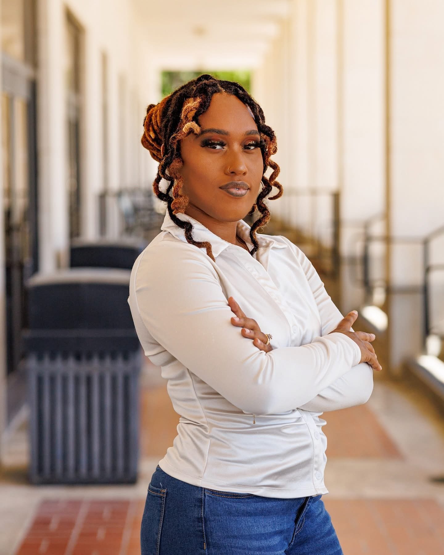 A woman with dreadlocks stands with arms crossed, wearing a white shirt and blue jeans, in a hallway with windows and a trash bin in the background.