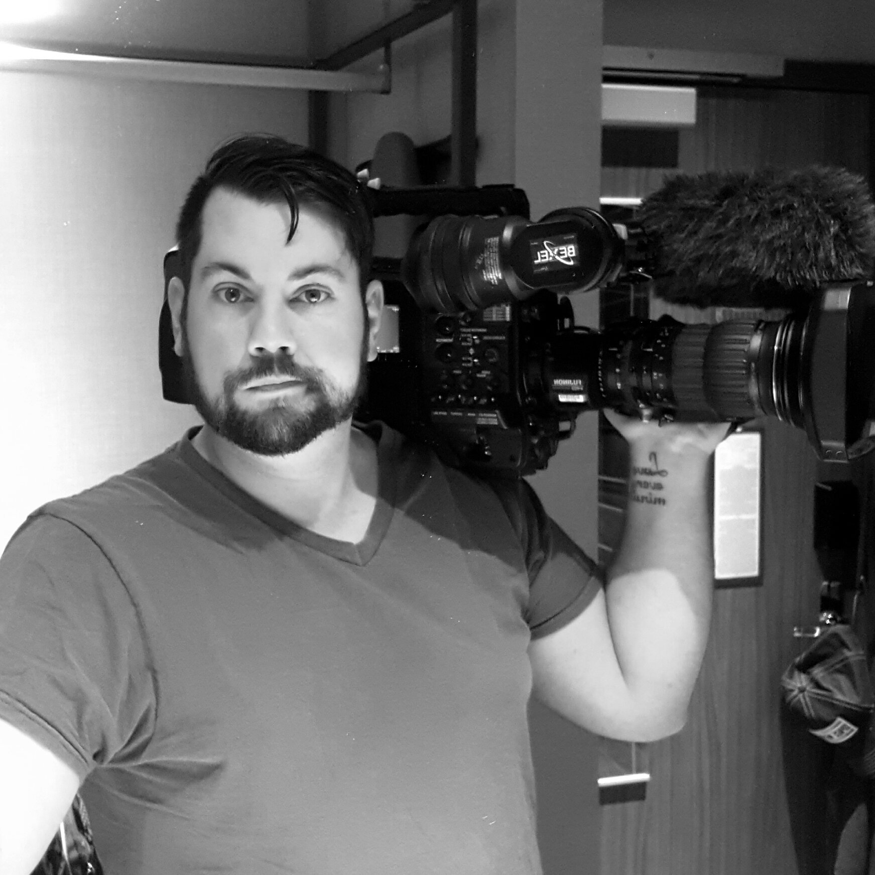 A man with dark hair and a beard holding a professional video camera on his shoulder, facing the camera, indoors.