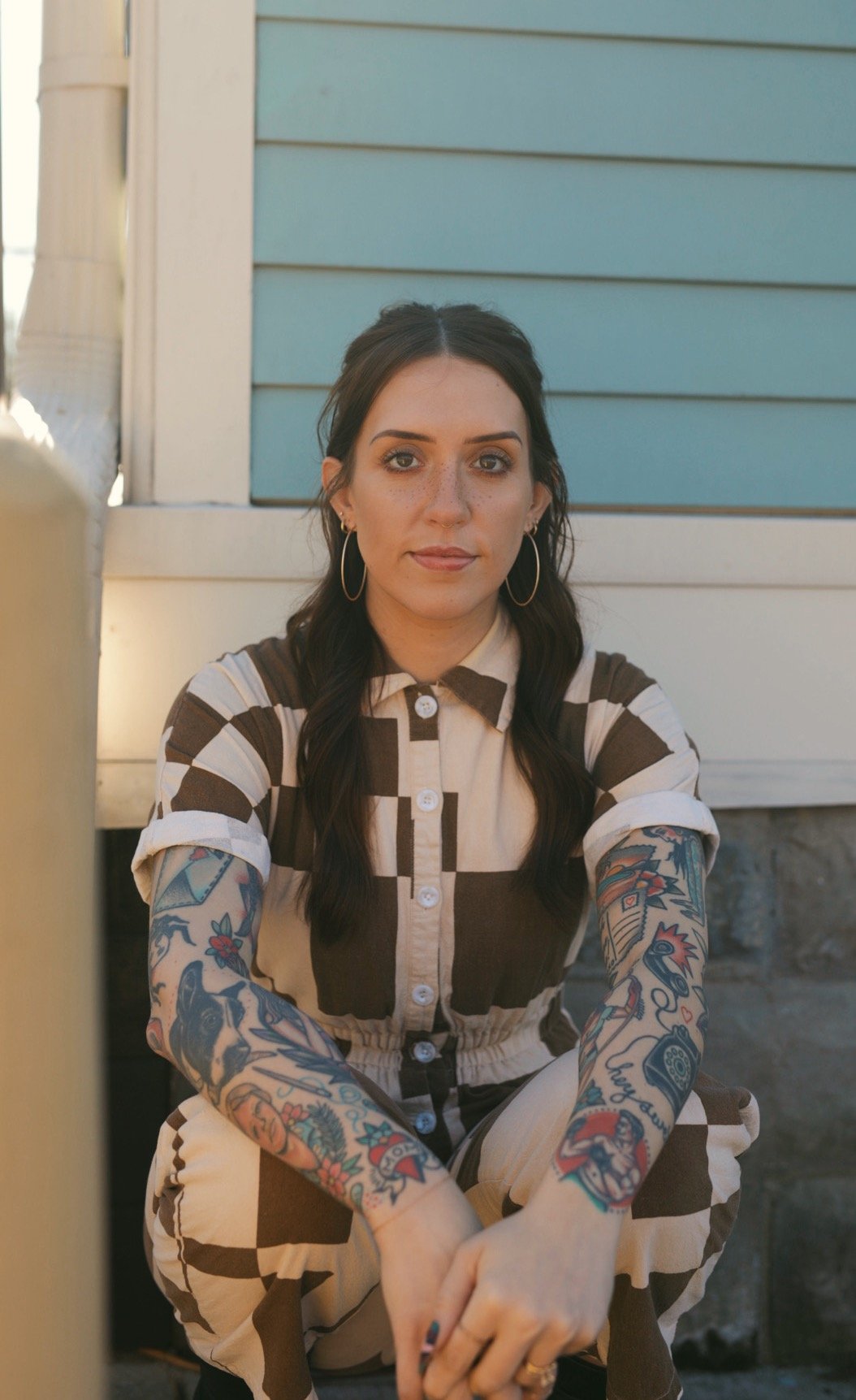A woman with dark hair, wearing large hoop earrings, a button-up dress with a brown and white checkered pattern, and has colorful tattoos on her arms, is sitting outdoors against a light blue house wall.