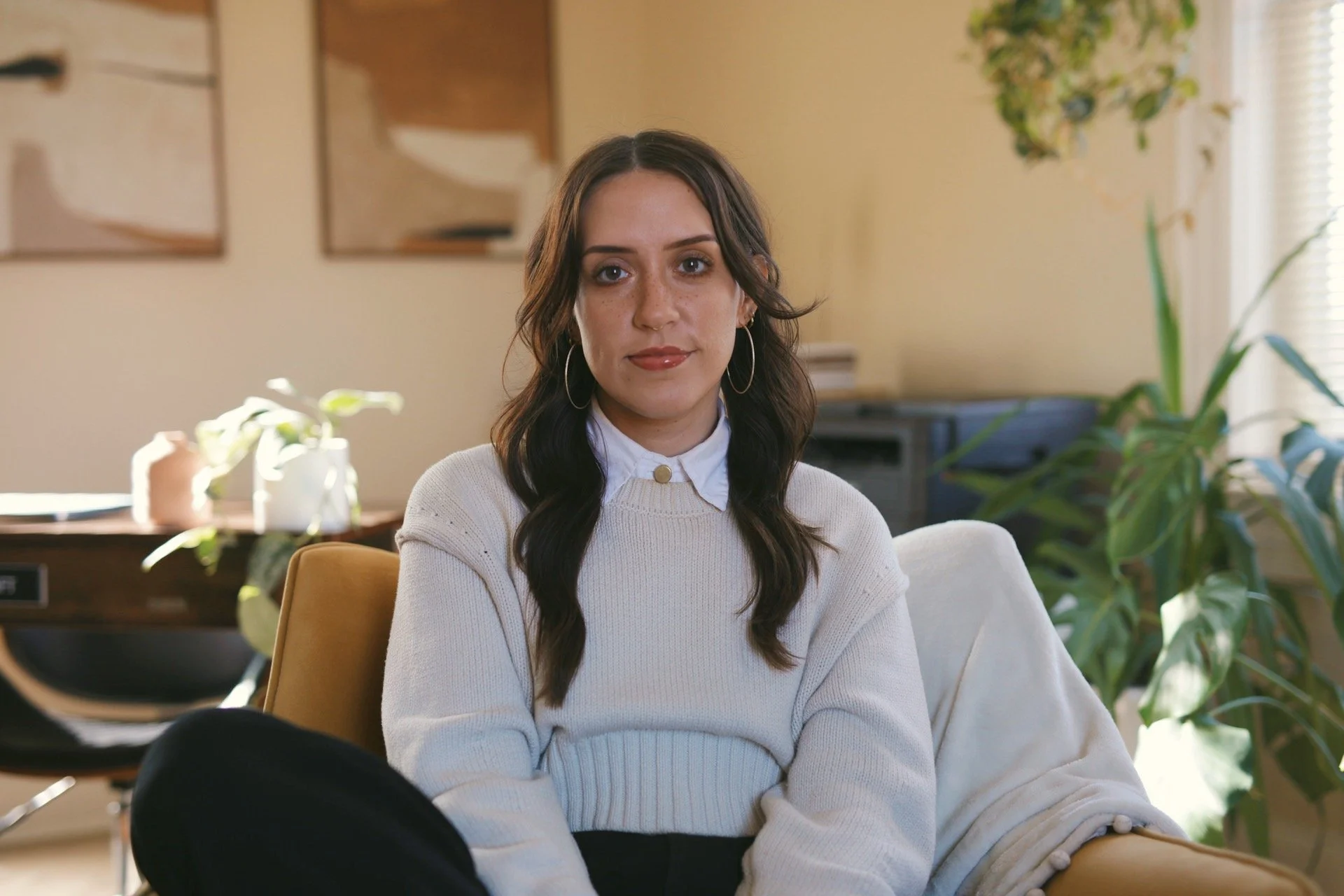 A woman with long, dark hair and hoop earrings, wearing a cream-colored sweater over a white collared shirt, sitting on a beige couch in a well-lit living room with plants and artwork in the background.
