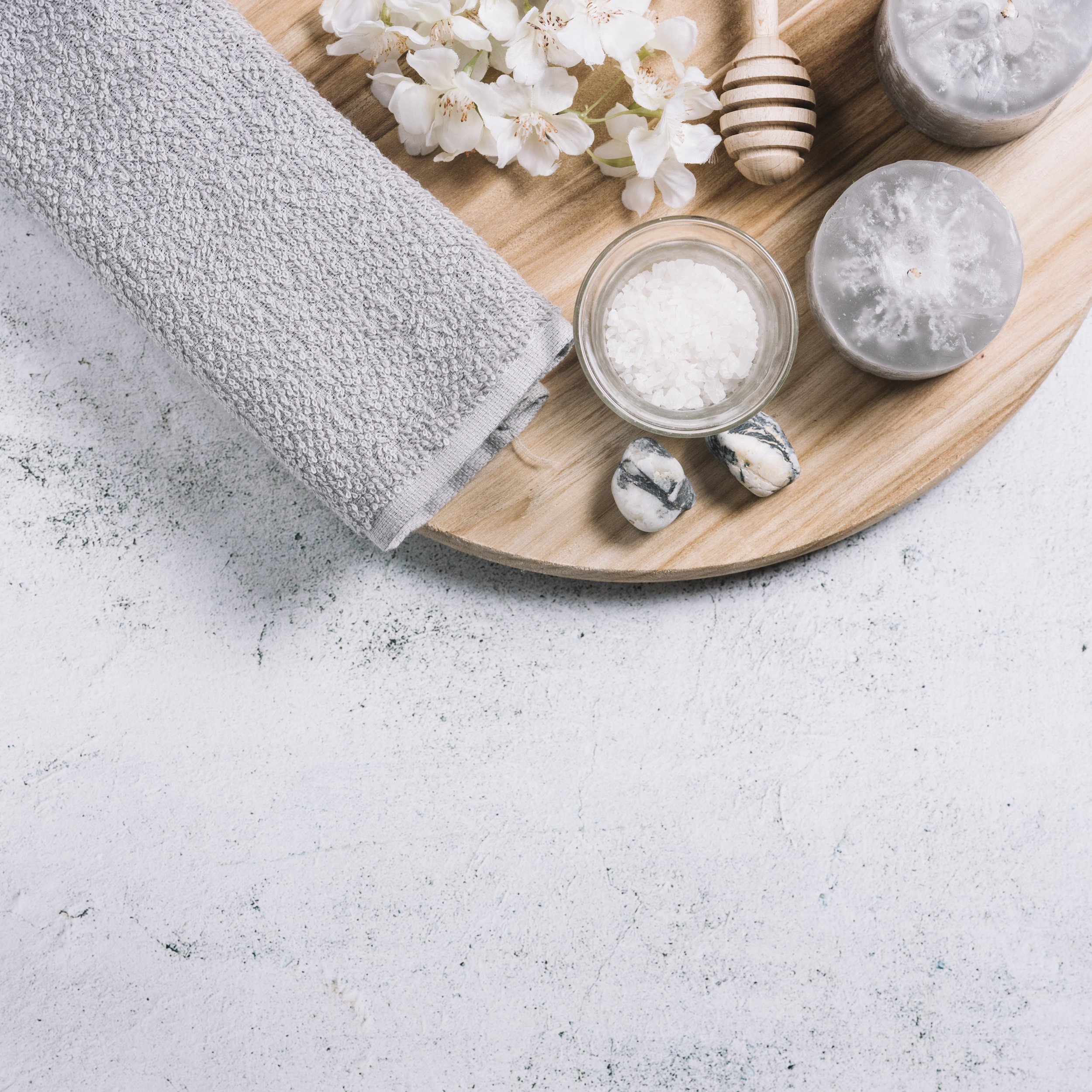 Spa setting with gray towel, white flowers, honey dipper, candles, salt, and stones on a round wooden tray.