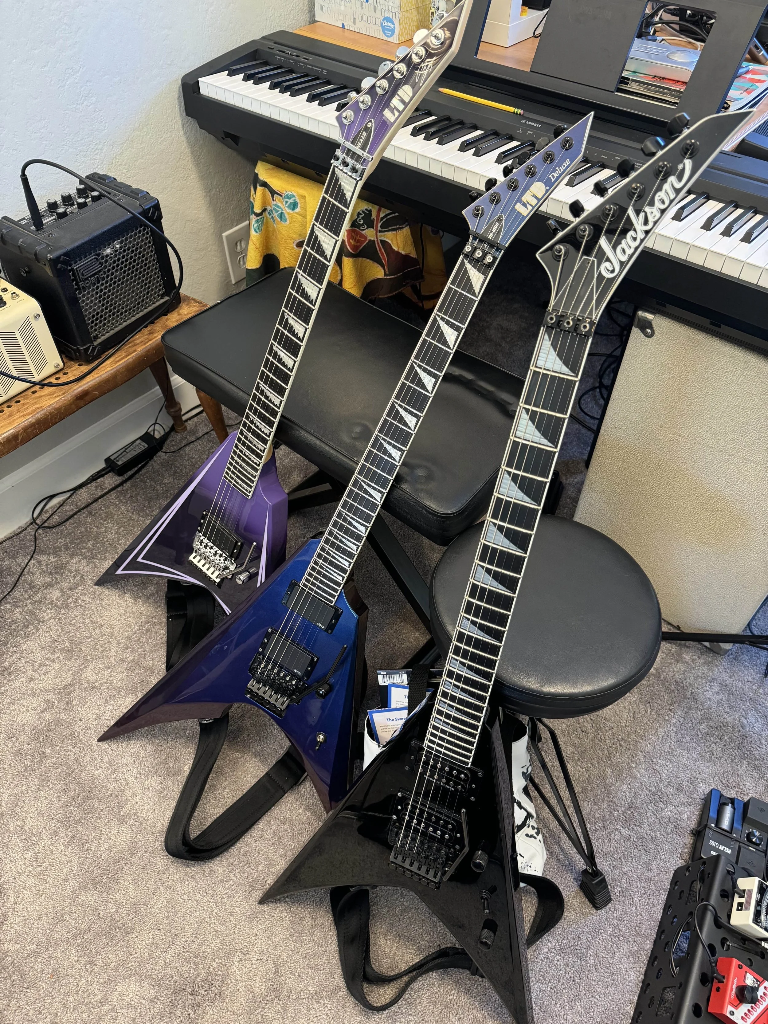 Three electric guitars with V-shaped bodies and dark metallic finishes, resting against a stool and a chair in a music room with a keyboard, amplifier, and music equipment.