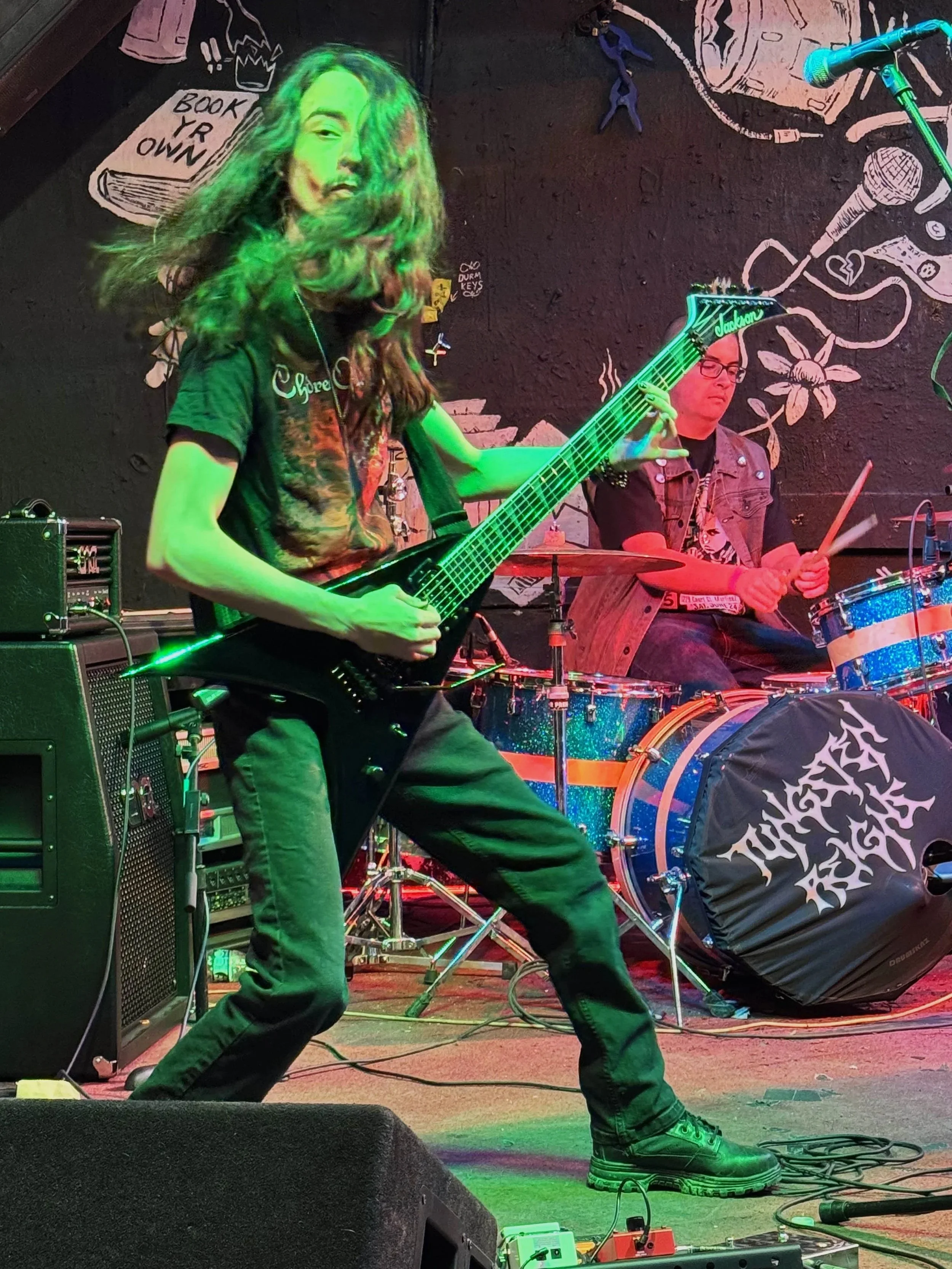 Aidan is playing the electric guitar on stage with long hair covering part of his face under green and red lighting.