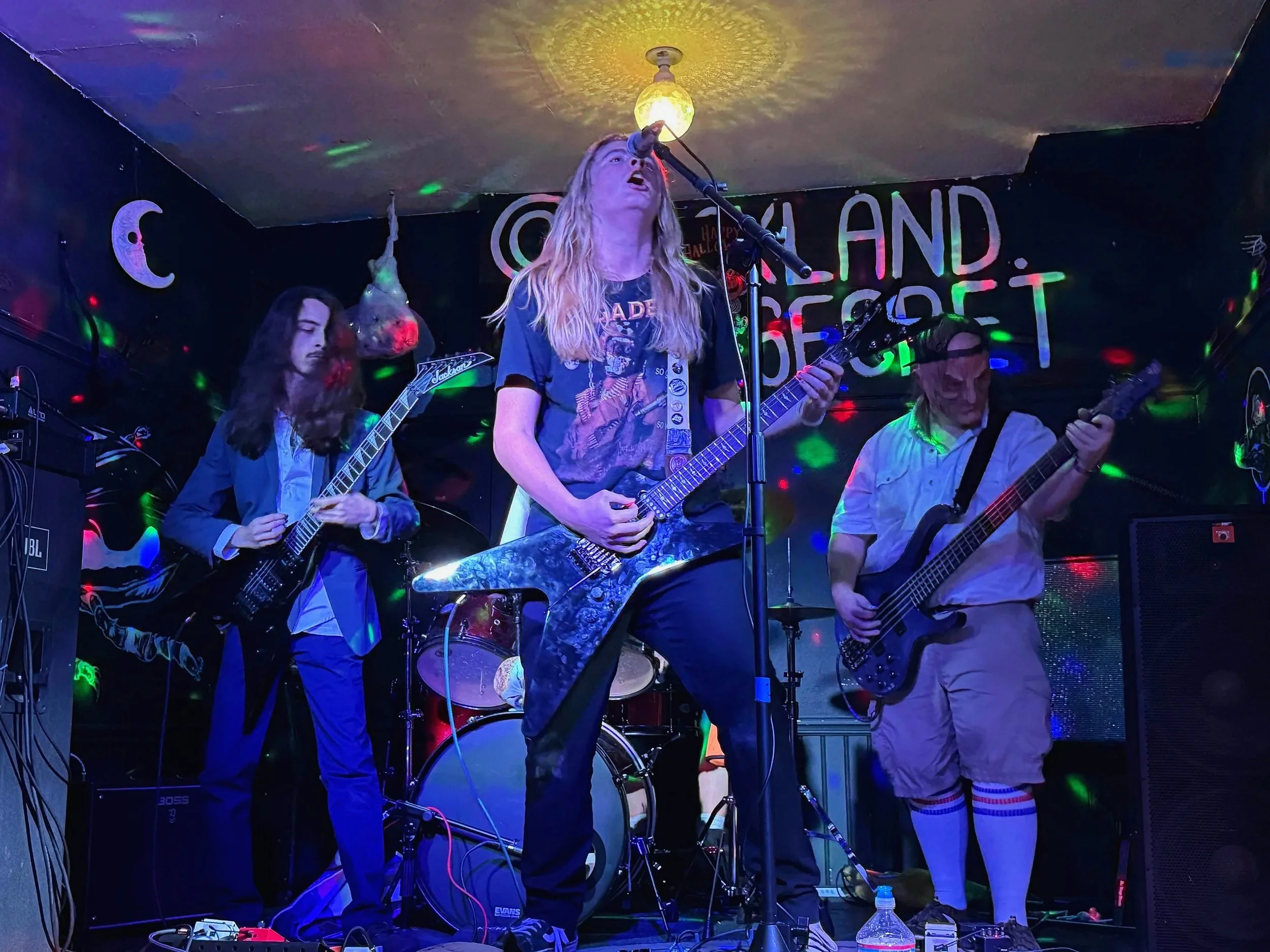 A musical band performing on stage with colorful lights. The lead singer is playing an electric guitar and singing into a microphone, while other band members are playing guitars and drums. The background has a banner with partial text 'ISLAND' and some decorations, including a crescent moon.
