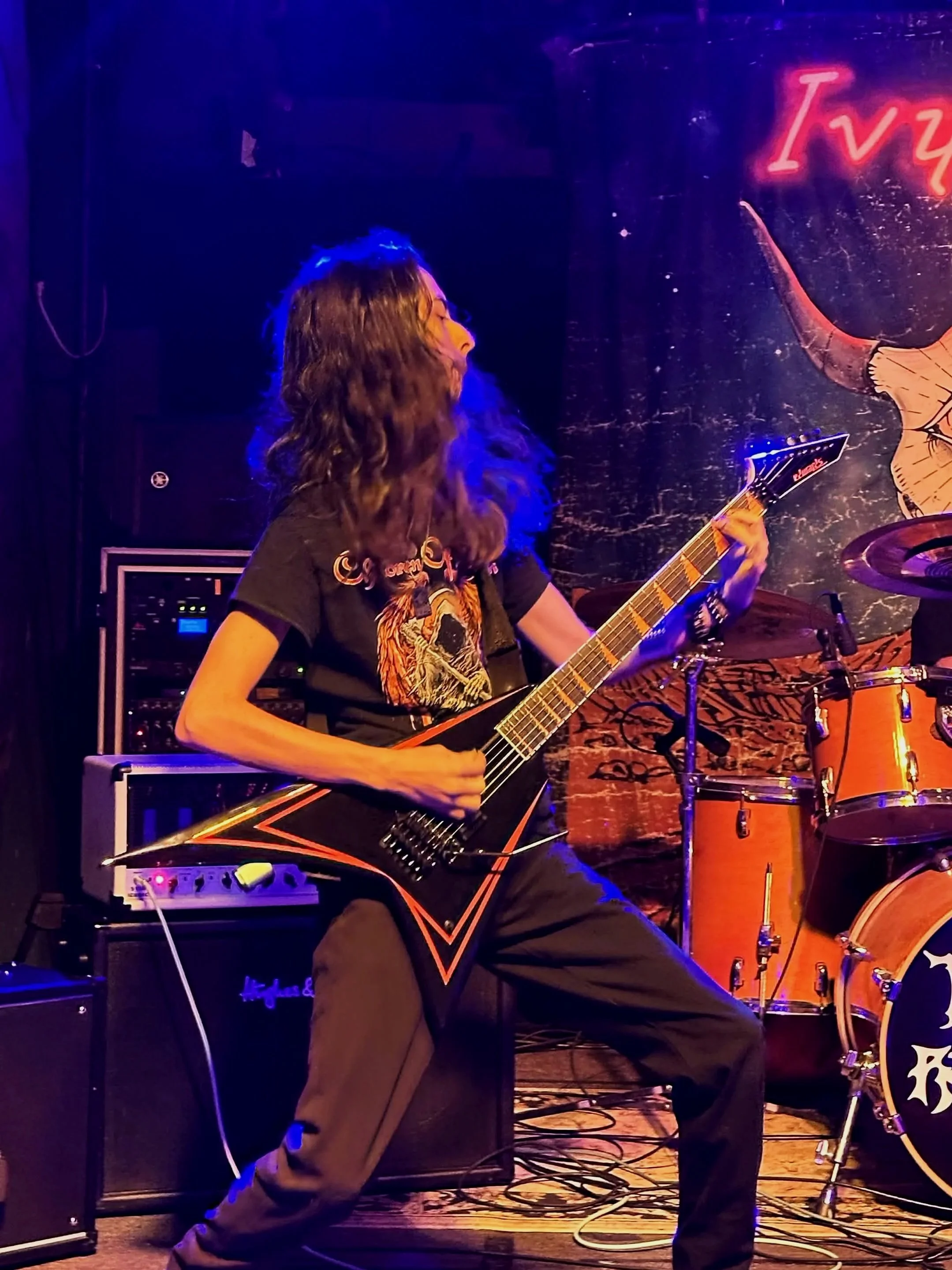 A person with long hair playing an electric guitar on stage, with musical equipment and drums in the background.