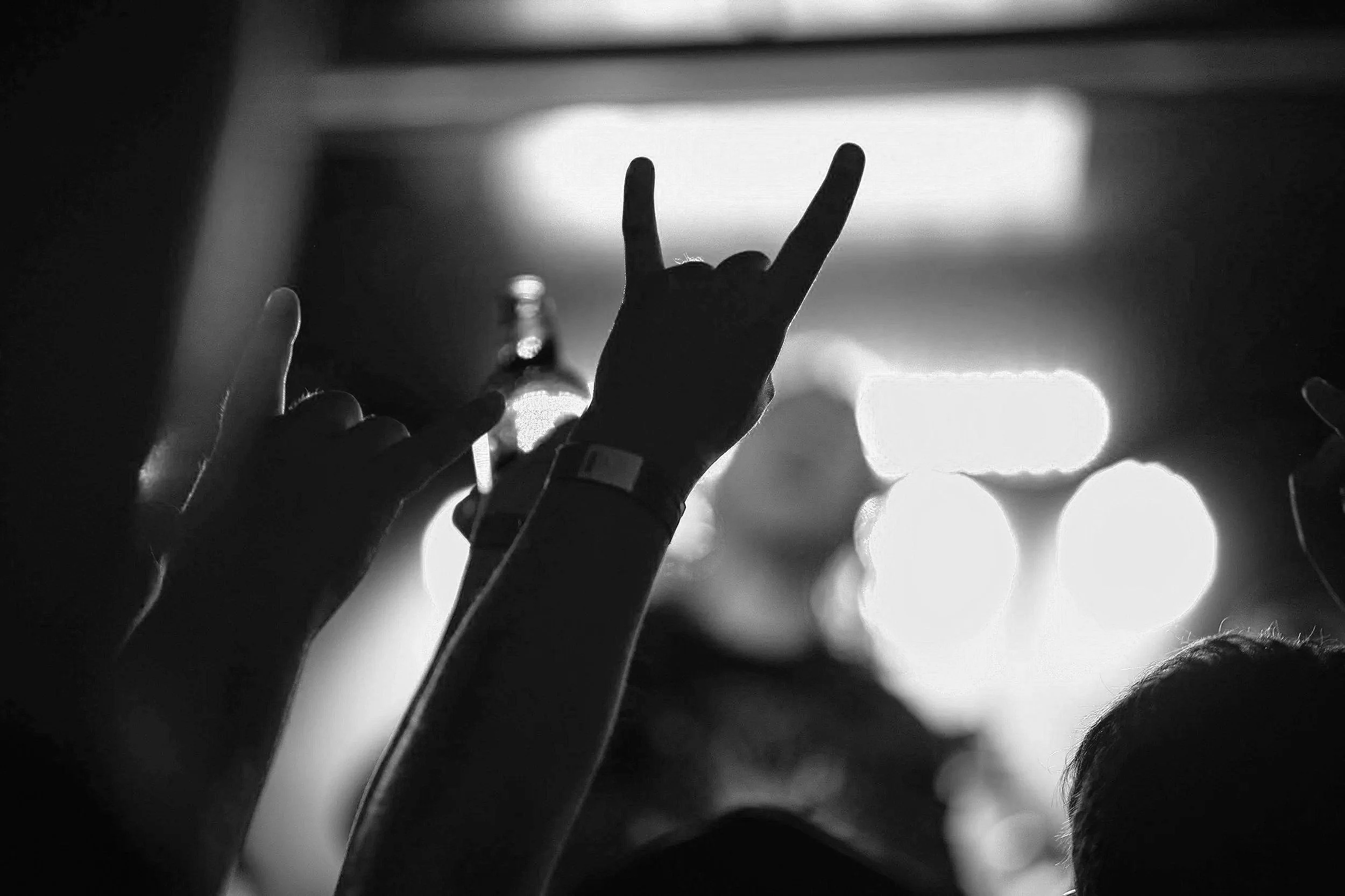 Black and white photo of people at a concert with hands raised, some making the 'rock on' gesture, in front of bright stage lights.
