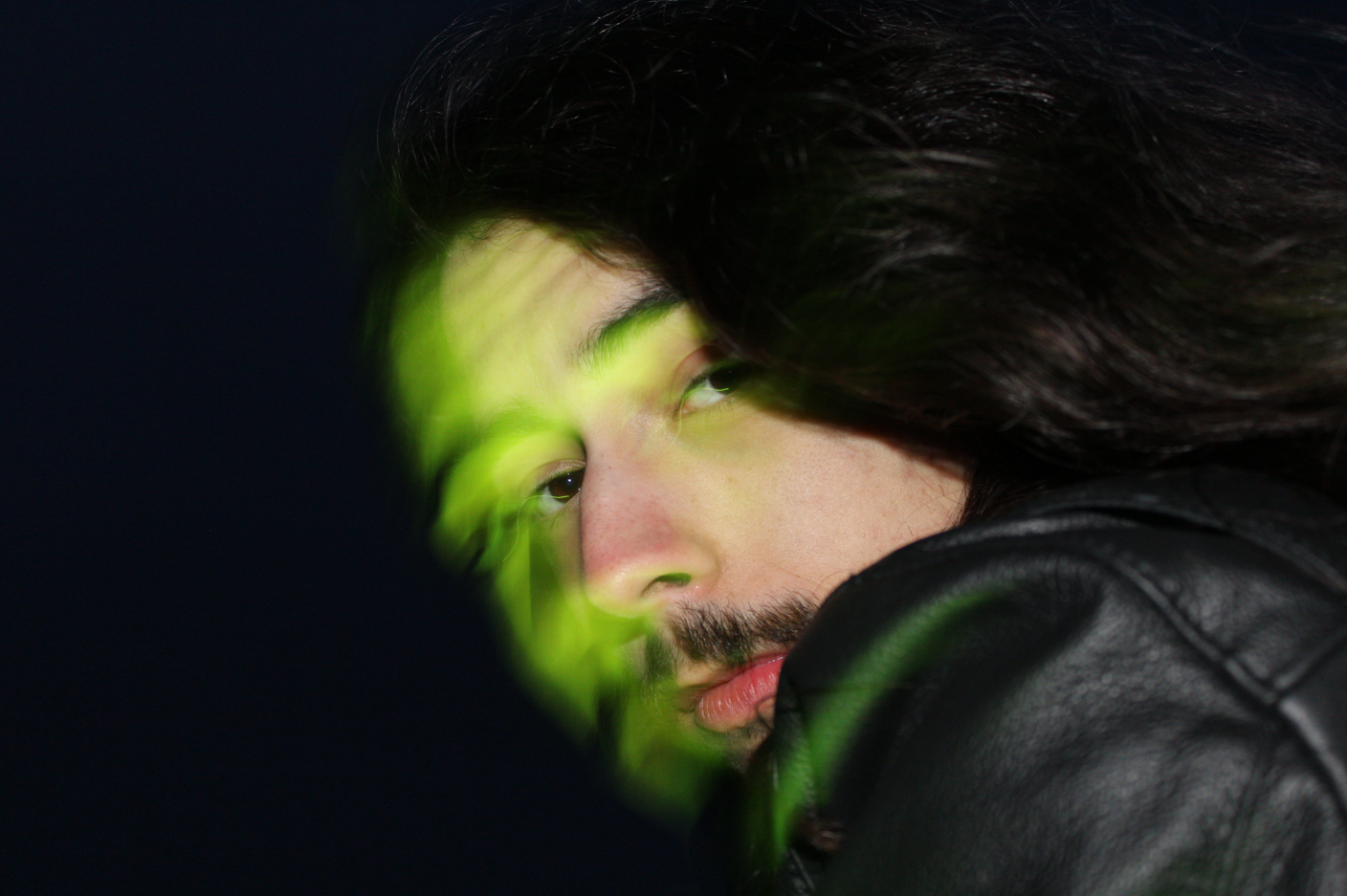 Close-up of a man's face with long dark hair and beard, lying down or leaning against a dark surface, partially obscured by green light reflections.
