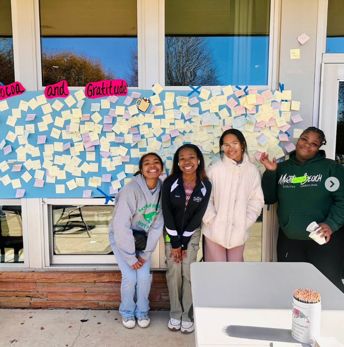 Four smiling girls stand in front of a blue wall decorated with numerous sticky notes, with the sign 'Cocoa and Gratitude' on top. A white table with a container of colored pencils is in the foreground.