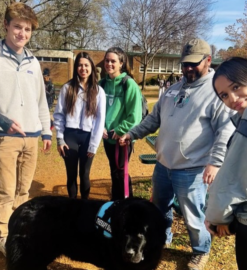 Group of five young people and a man standing outdoors with a black dog in front of them. The scene takes place on a sunny day, with trees and a building in the background. The dog is lying on the ground, wearing a harness, and the group appears to be enjoying a casual gathering in a park or schoolyard.