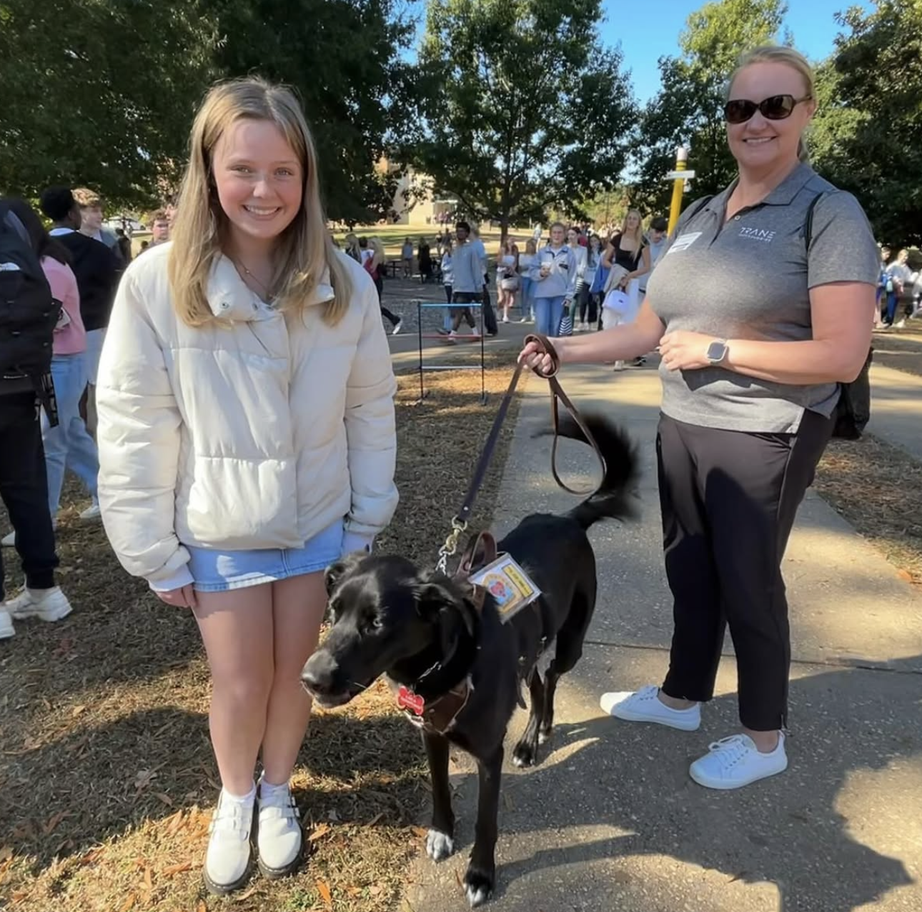 A young blonde girl smiling and standing next to a black service dog on a leash. The dog wears a vest and is looking at the camera. An older woman with sunglasses and a gray polo shirt stands beside them, holding the leash. There are other people and trees in the background on a sunny day.