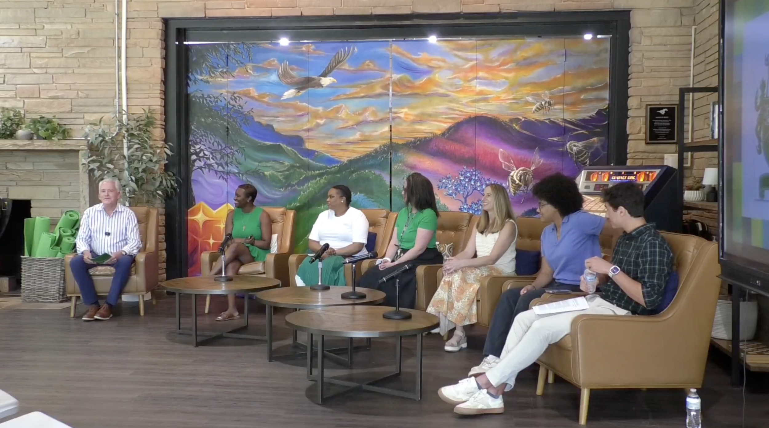 Panel discussion with seven people seated on tan leather chairs in a room with a colorful mountain landscape mural behind them. Four women and three men are present, some with microphones. A game of air hockey is visible in the background.