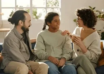 Three people sitting on a couch having a conversation, with large windows in the background.