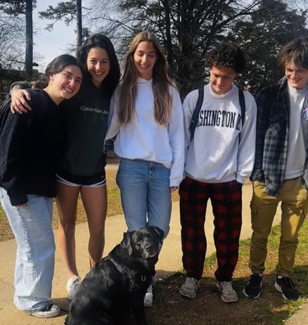 Group of five teenagers standing outdoors with a black dog sitting in front of them, all smiling, with trees and a walk path in the background.