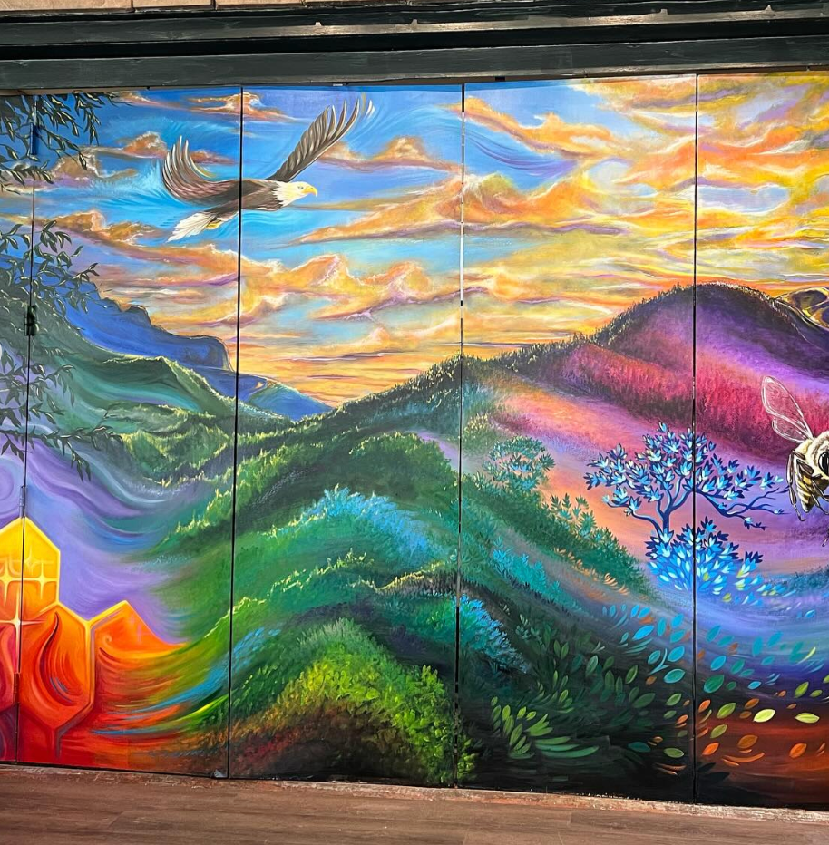Colorful mural depicting a landscape with mountains, trees, and clouds at sunset or sunrise. An eagle is flying in the sky, and a bee is near the bottom right corner.