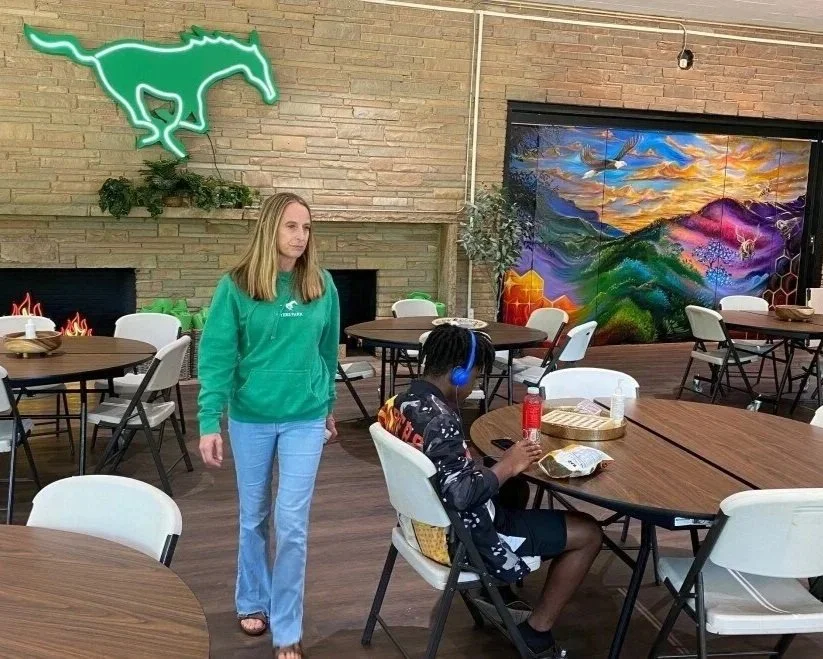A woman in a green hoodie and jeans stands in a room with several round tables and white chairs. A person with headphones sits at a table eating, with a fire-shaped decoration on a brick wall and colorful mural in the background.