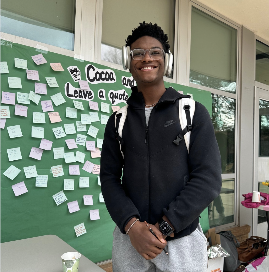A smiling young man wearing glasses, headphones, a black Nike jacket, and a white backpack standing in front of a green bulletin board with notes, in a room with large windows.
