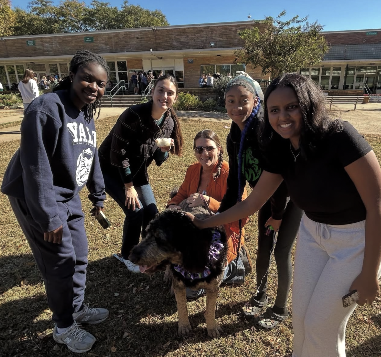 Group of five women and a large dog outside on grass in front of a school building. They are smiling and petting the dog, which is wearing a bandana. The women are dressed casually, and the weather appears sunny.