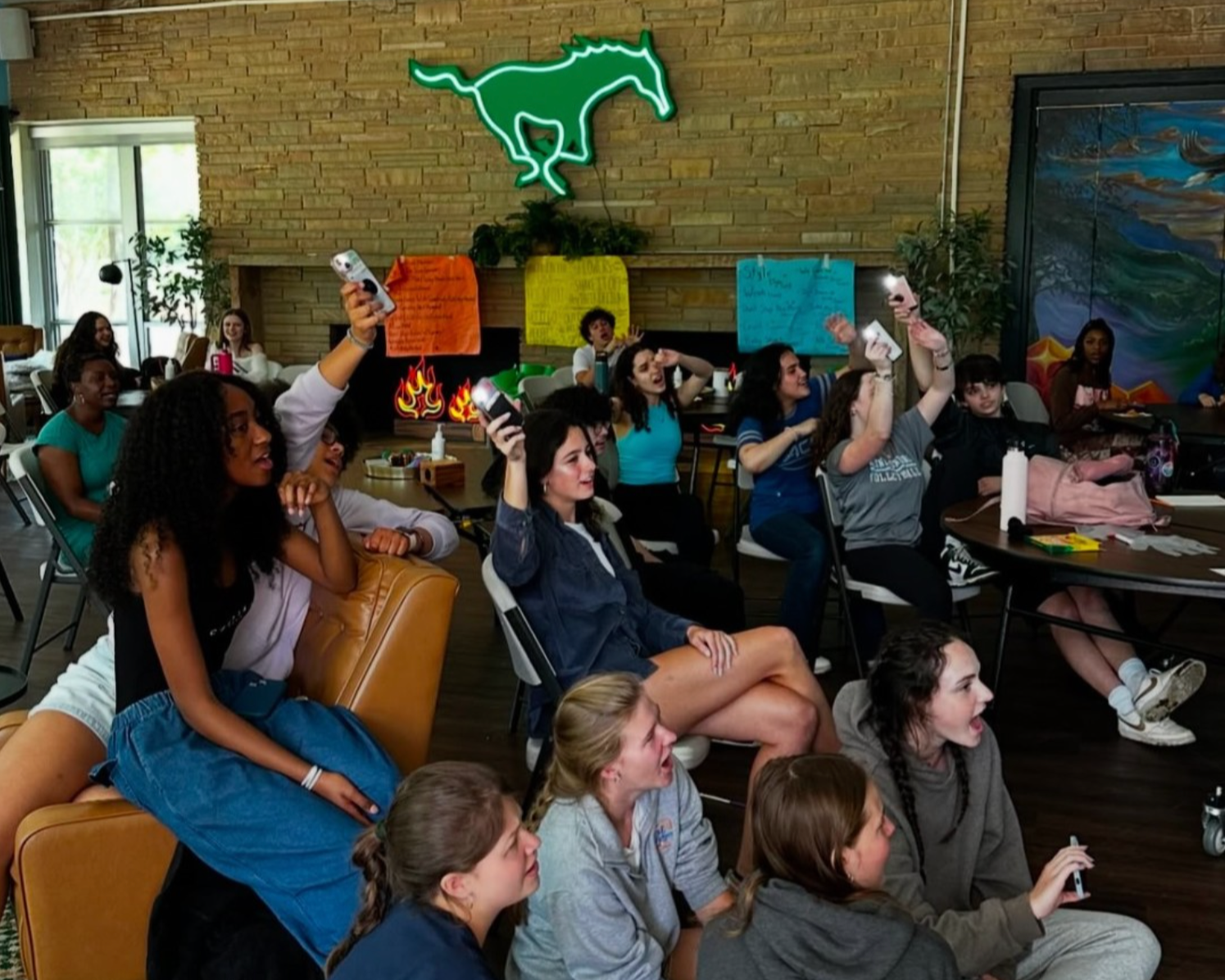 Group of young people attending a classroom or workshop, some raising hands, some sitting and listening, with colorful posters and a neon dinosaur sign on the brick wall.