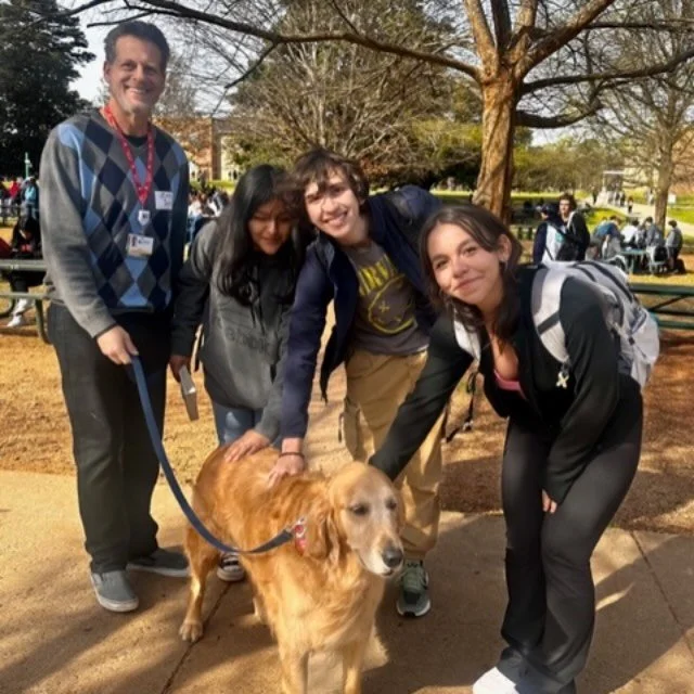 Four people smiling and petting a golden retriever dog outdoors during daytime with trees and other people in the background.