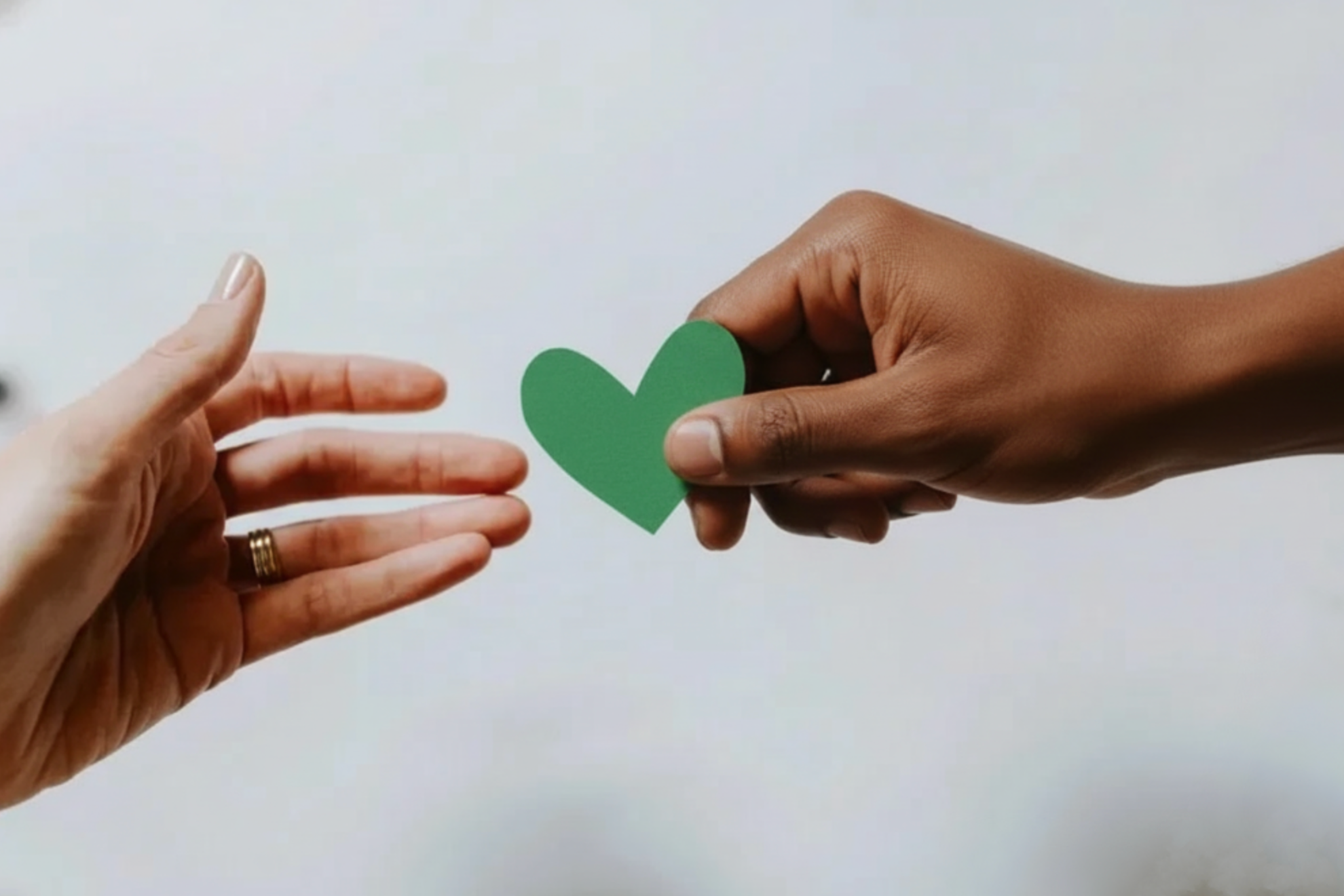 One person is handing a green paper heart to another person's outstretched hand against a plain background.