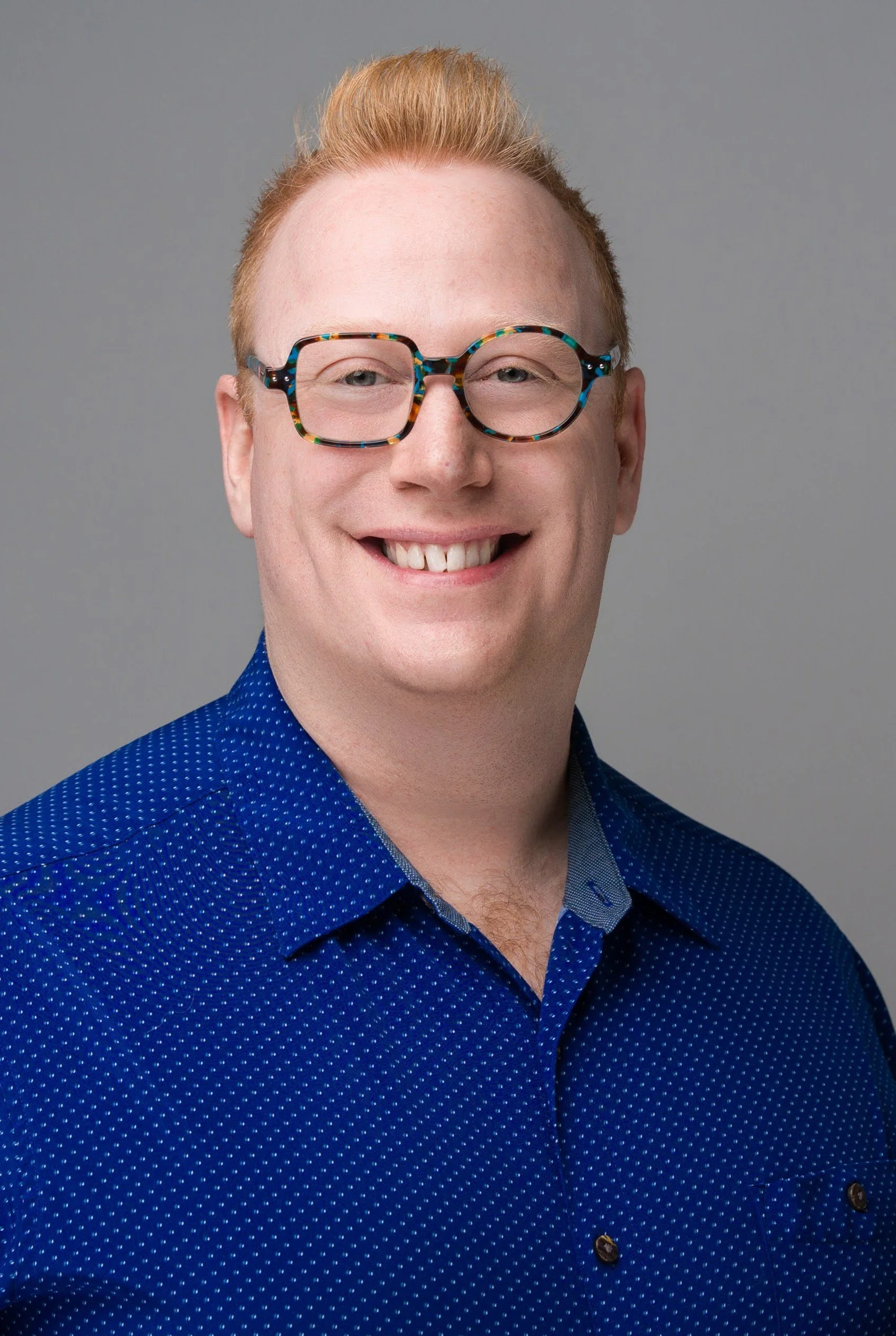 A smiling man with short red hair and glasses with a colorful frame, wearing a blue patterned shirt, against a plain gray background.