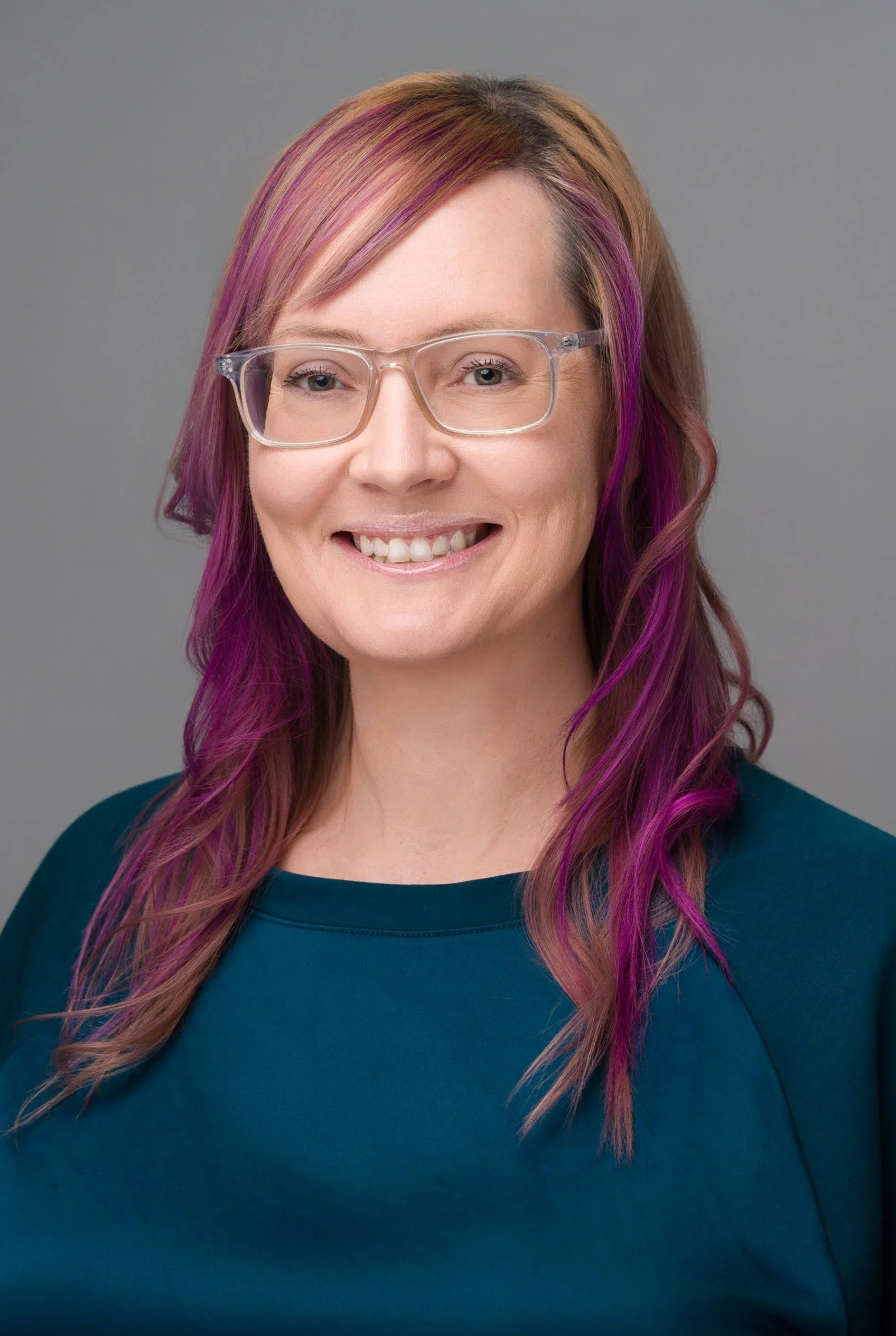 A woman with glasses and long, wavy hair dyed pink and purple, smiling at the camera, wearing a teal top, against a gray background.