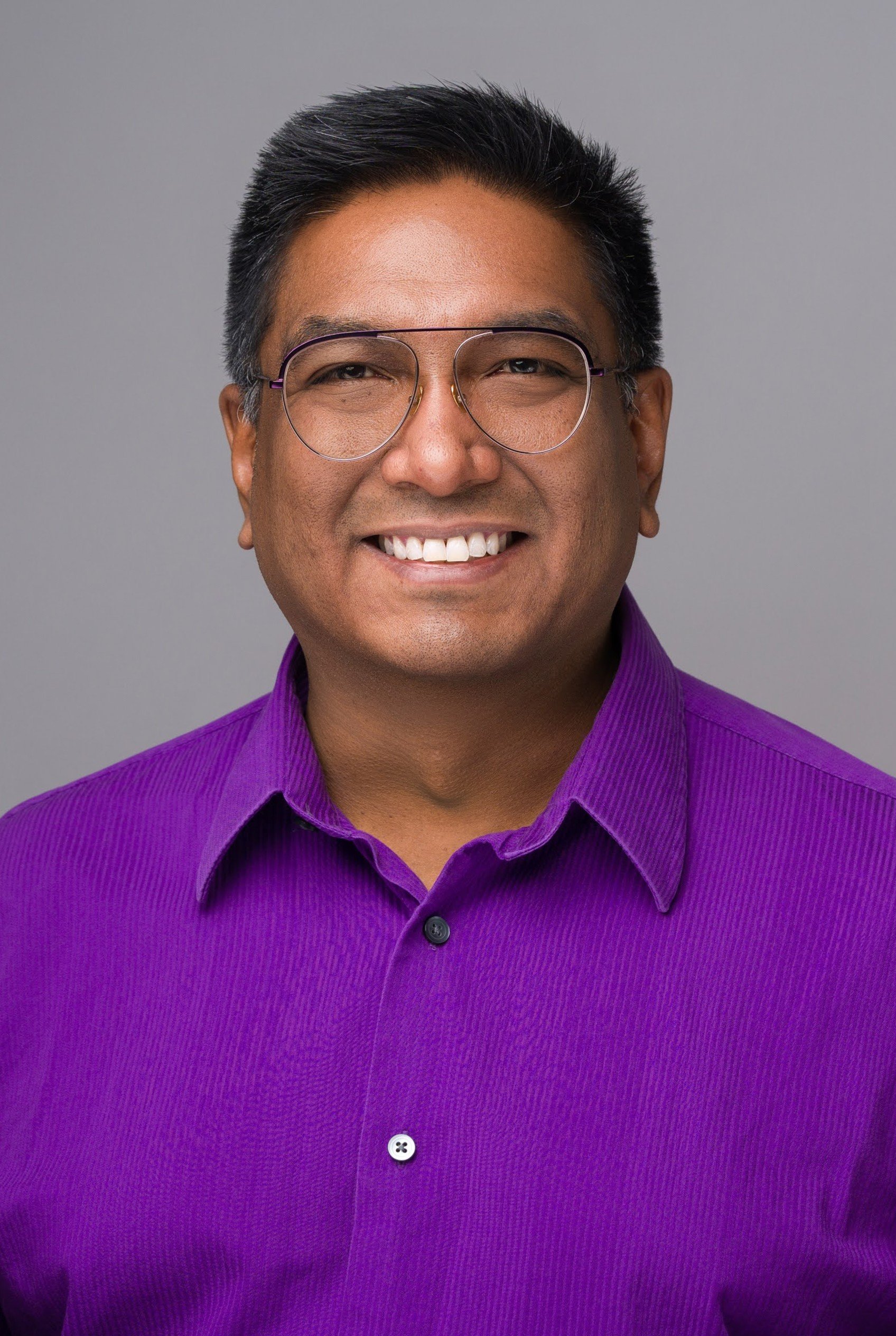 Headshot of a smiling man wearing glasses and a purple shirt against a neutral background.