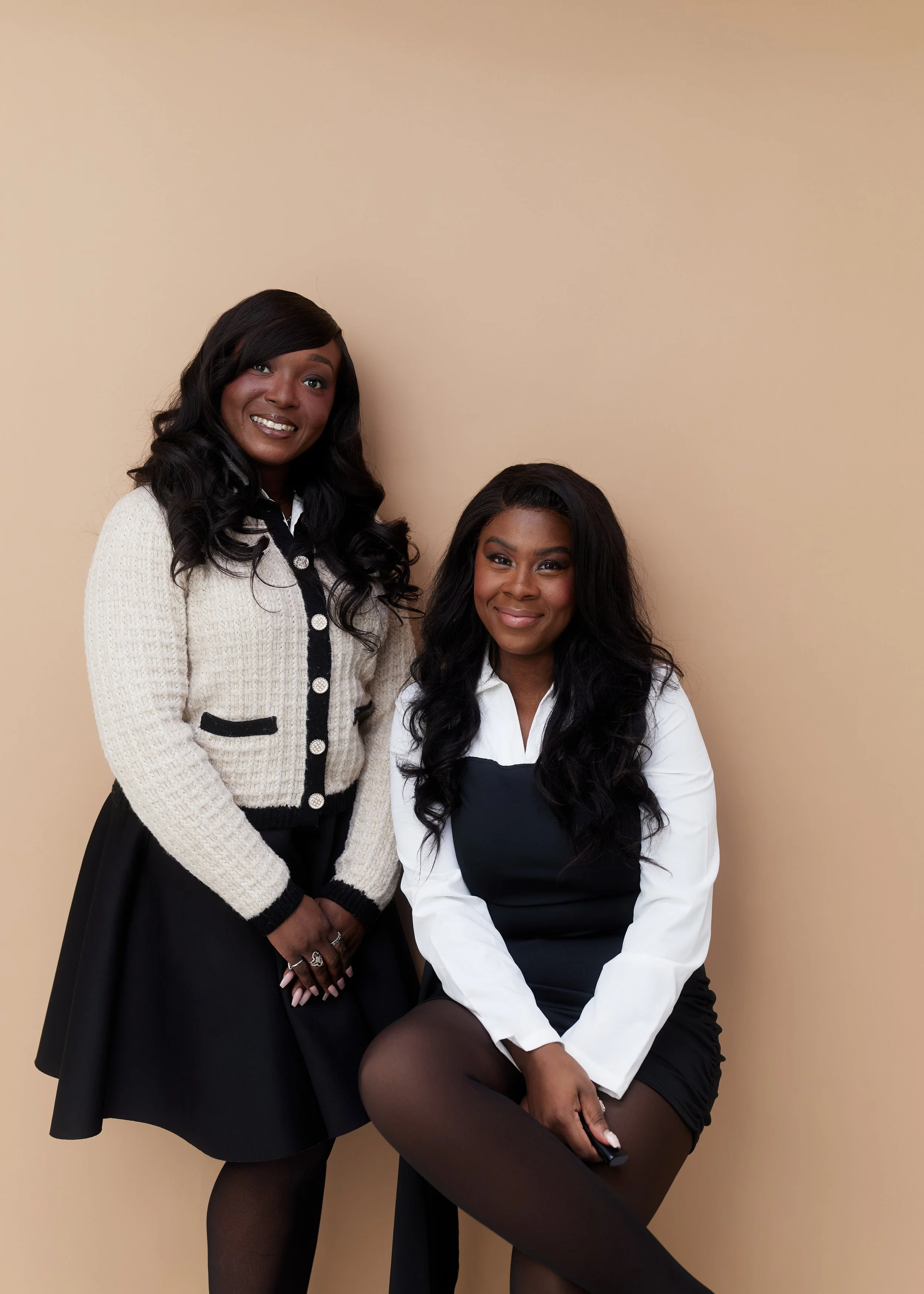 Two women with long dark hair and dark skin, smiling, posing against a beige background. One woman is standing, wearing a cream-colored jacket with black trim and a black skirt. The other woman is sitting, wearing a white blouse with a black dress over it and black tights.