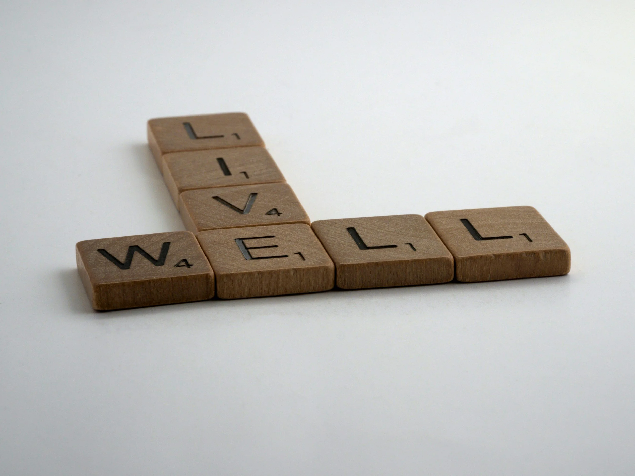 Wooden Scrabble tiles spelling out the words 'LIVE' vertically and 'WELL' horizontally on a white surface.