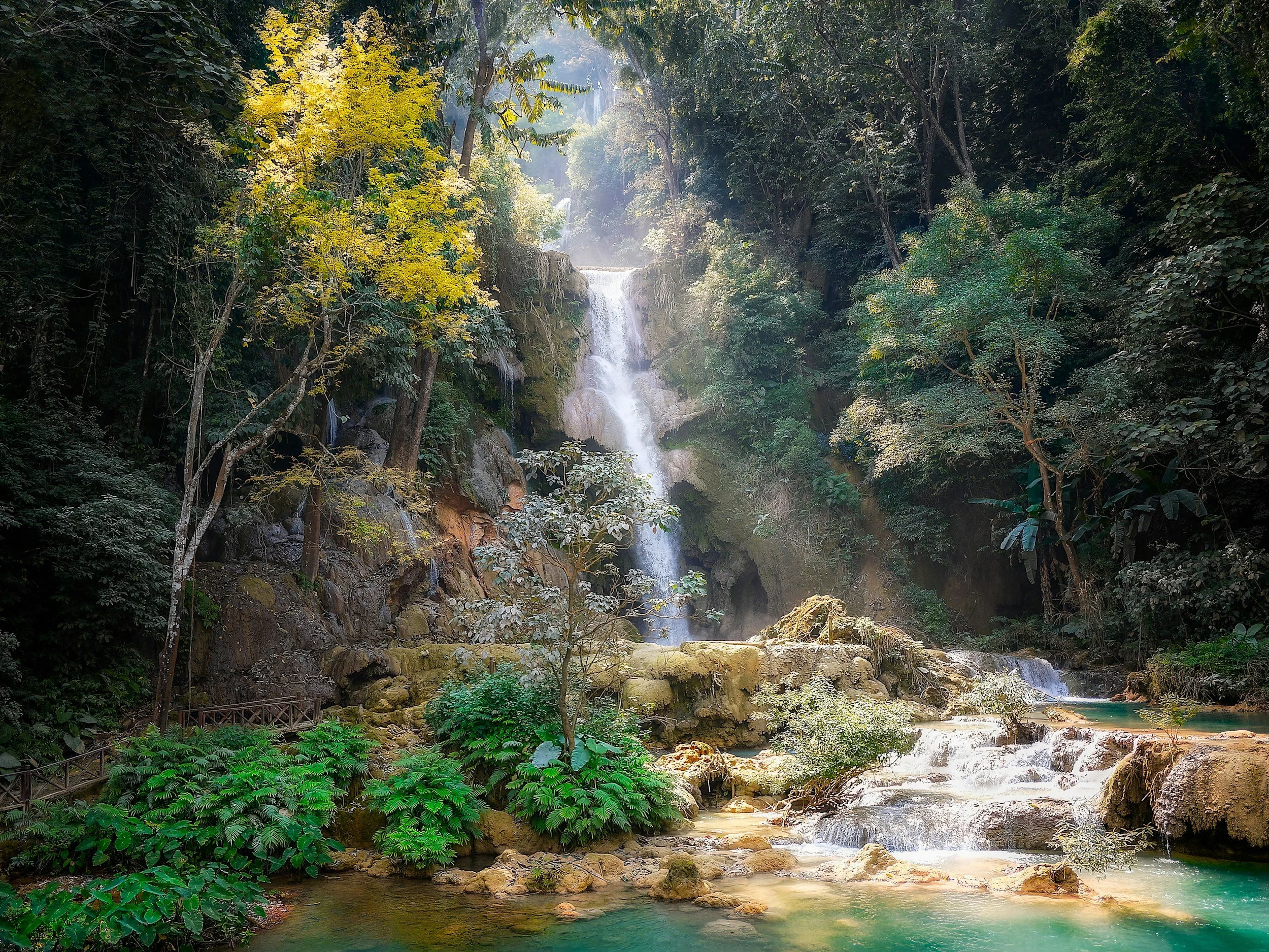 A multi-tiered waterfall flowing into a turquoise pool surrounded by lush green trees and foliage.