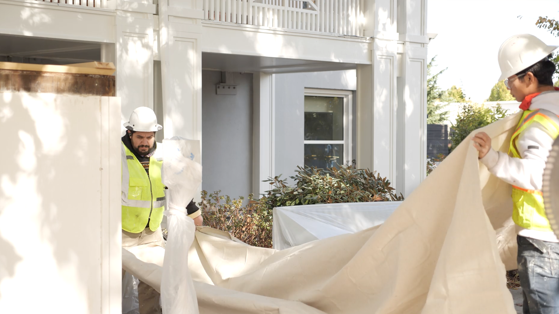 Two construction workers, a man and a woman, wearing white safety helmets and yellow reflective vests, working together on a building project outside a white house. The woman is holding a large piece of protective fabric, and the man appears to be preparing or adjusting something.