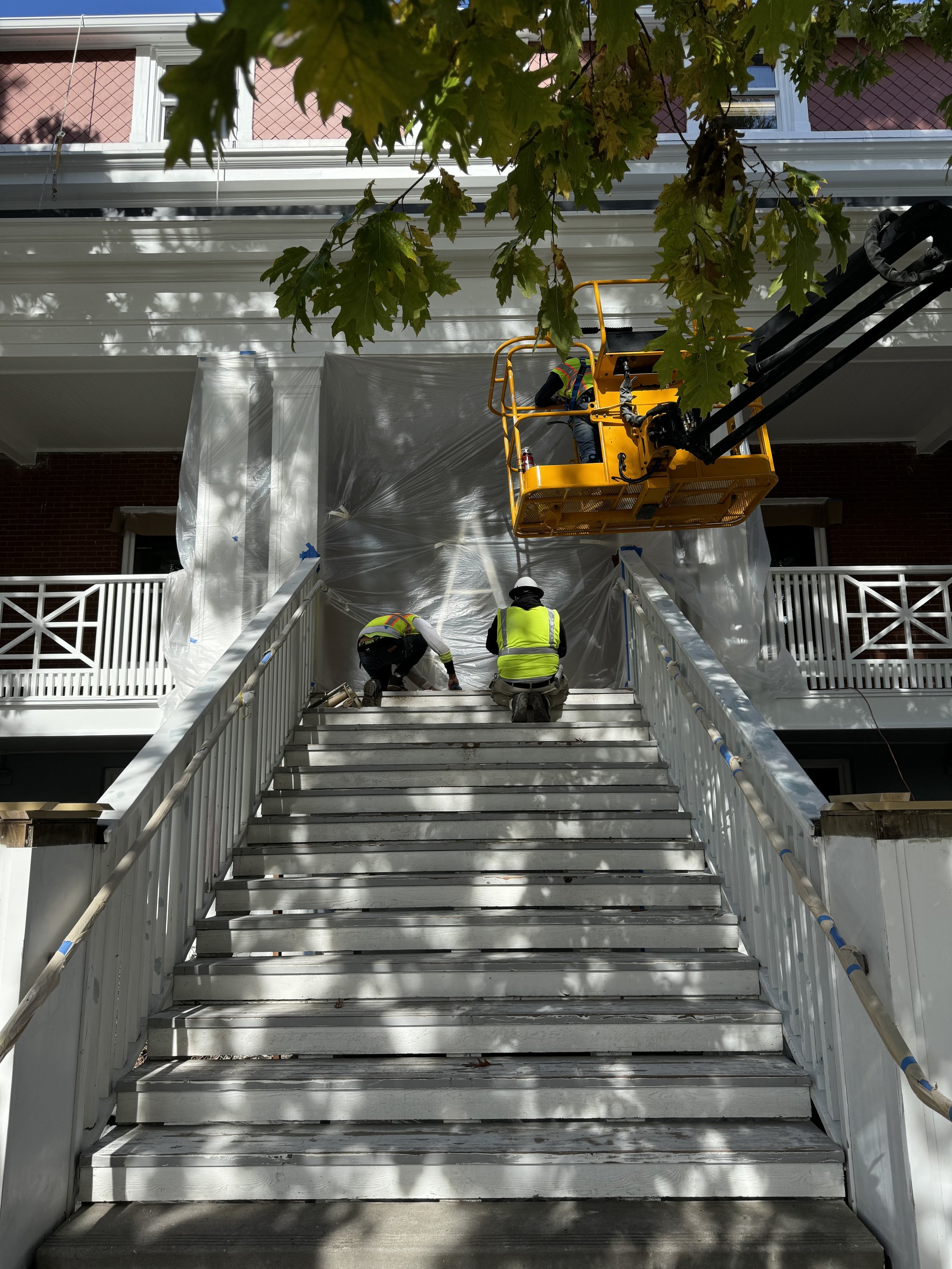 Construction workers repairing or painting outside stairs of a building, with a yellow lift and plastic sheeting covering parts of the building, in a residential area with trees overhead.