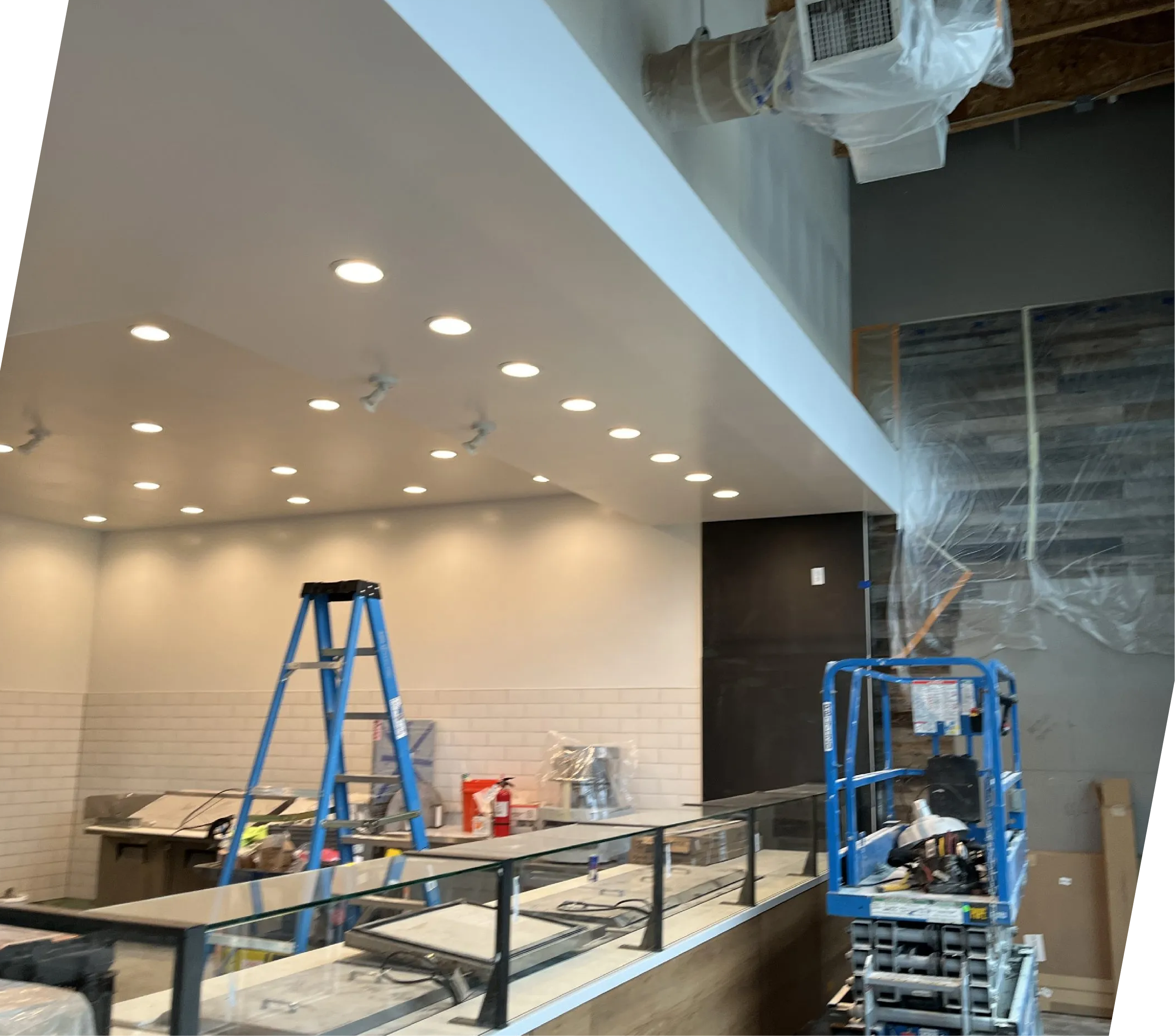 Construction interior with ladders, scaffolding, tools, and supplies on metal and wooden shelves, ceiling lights, and partially finished walls.