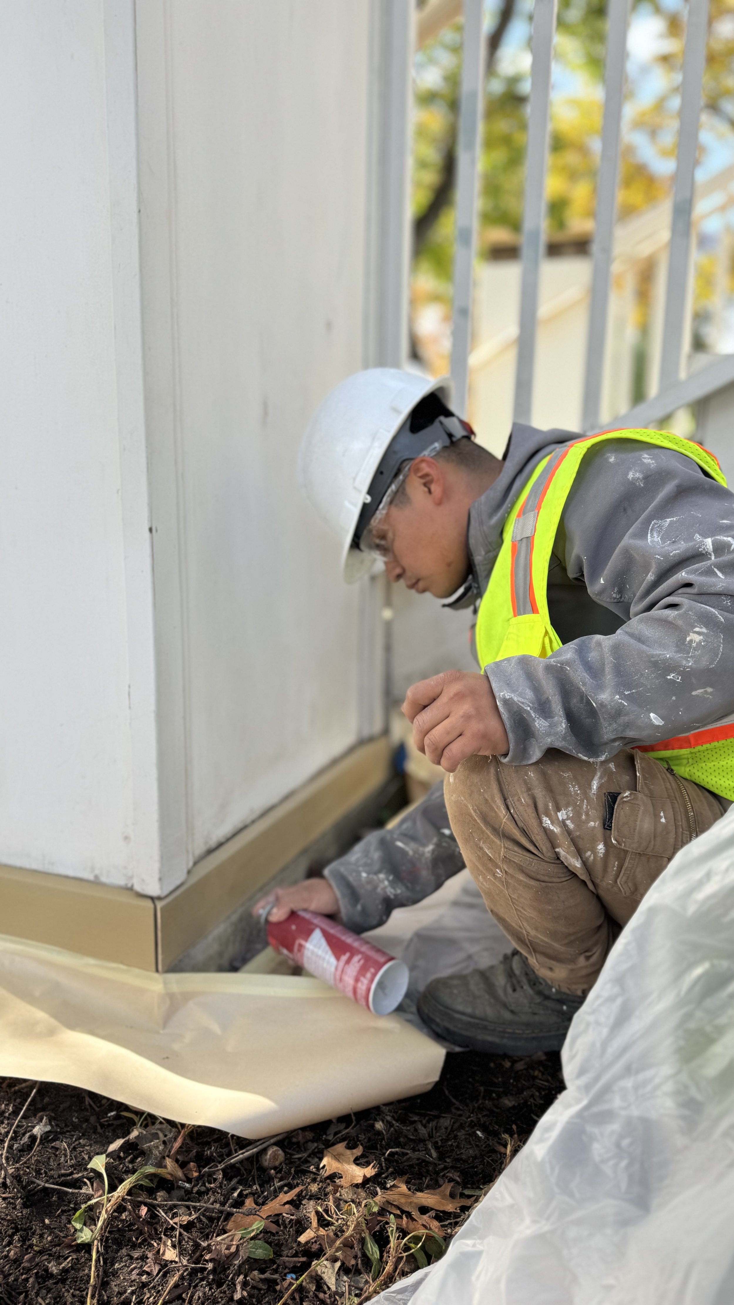 A construction worker wearing a white hard hat, safety vest, and paint-smudged clothes is kneeling on the ground, applying a sealant around the base of a white building with a tool in his hand. There is garden soil with some fallen leaves and a plastic sheet covering the ground nearby.