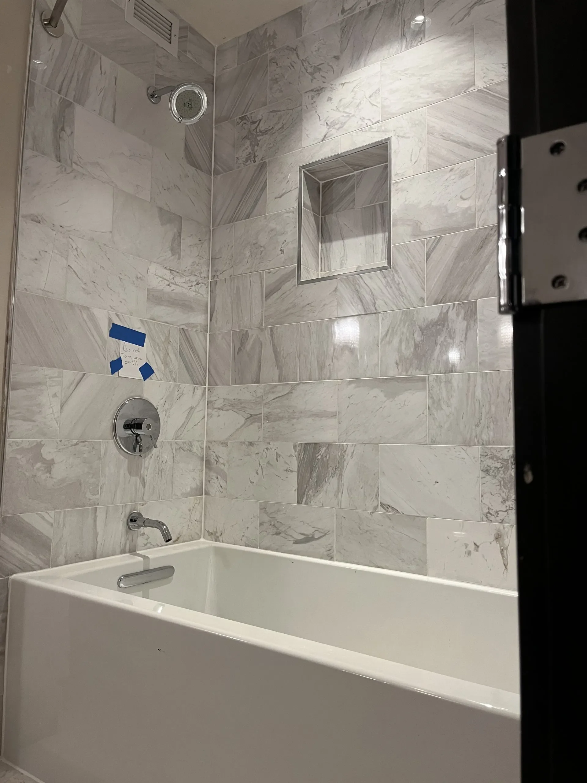Bathroom shower area with marble tile walls, a showerhead, a built-in niche, faucet, and bathtub, with a note that says 'Do not turn water on!!!' taped on the tile.